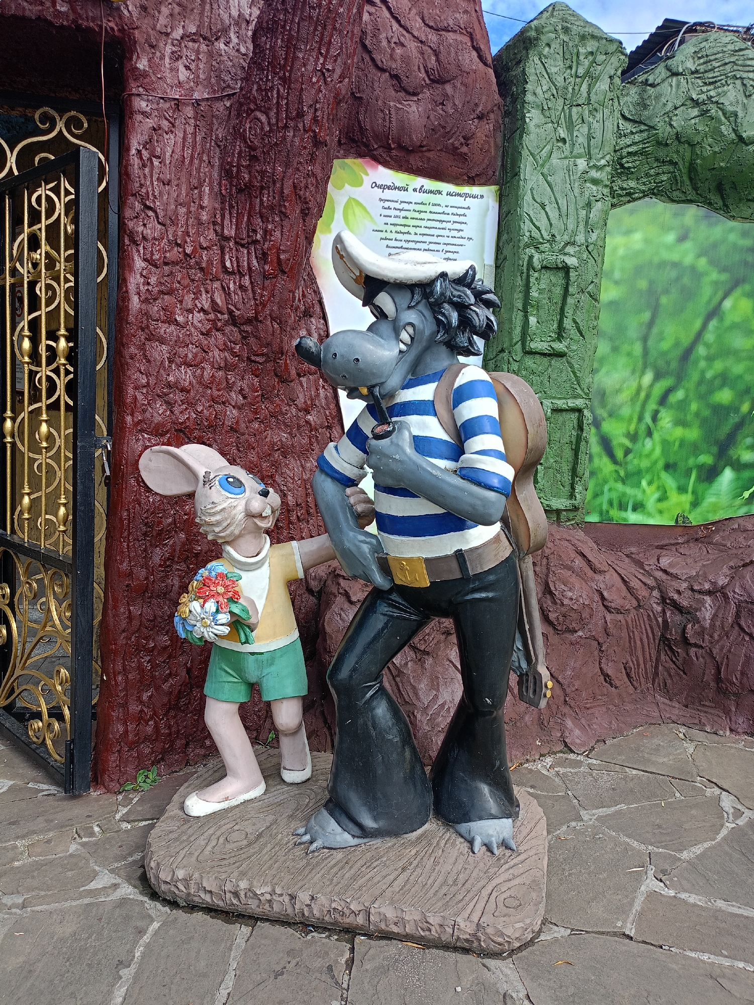A statue depicting characters from the Soviet animated series "Nu, pogodi!" is located in the city of Grozny, Russia. The statue features the wolf character, dressed in a sailor suit with a striped shirt and bell-bottom pants, holding a pipe and a guitar. Beside him stands the hare character, wearing a yellow shirt and green shorts, holding a bouquet of colorful flowers. They are positioned in front of a stylized tree trunk and a green archway that displays a nature-themed mural. The ground is paved with stone tiles.