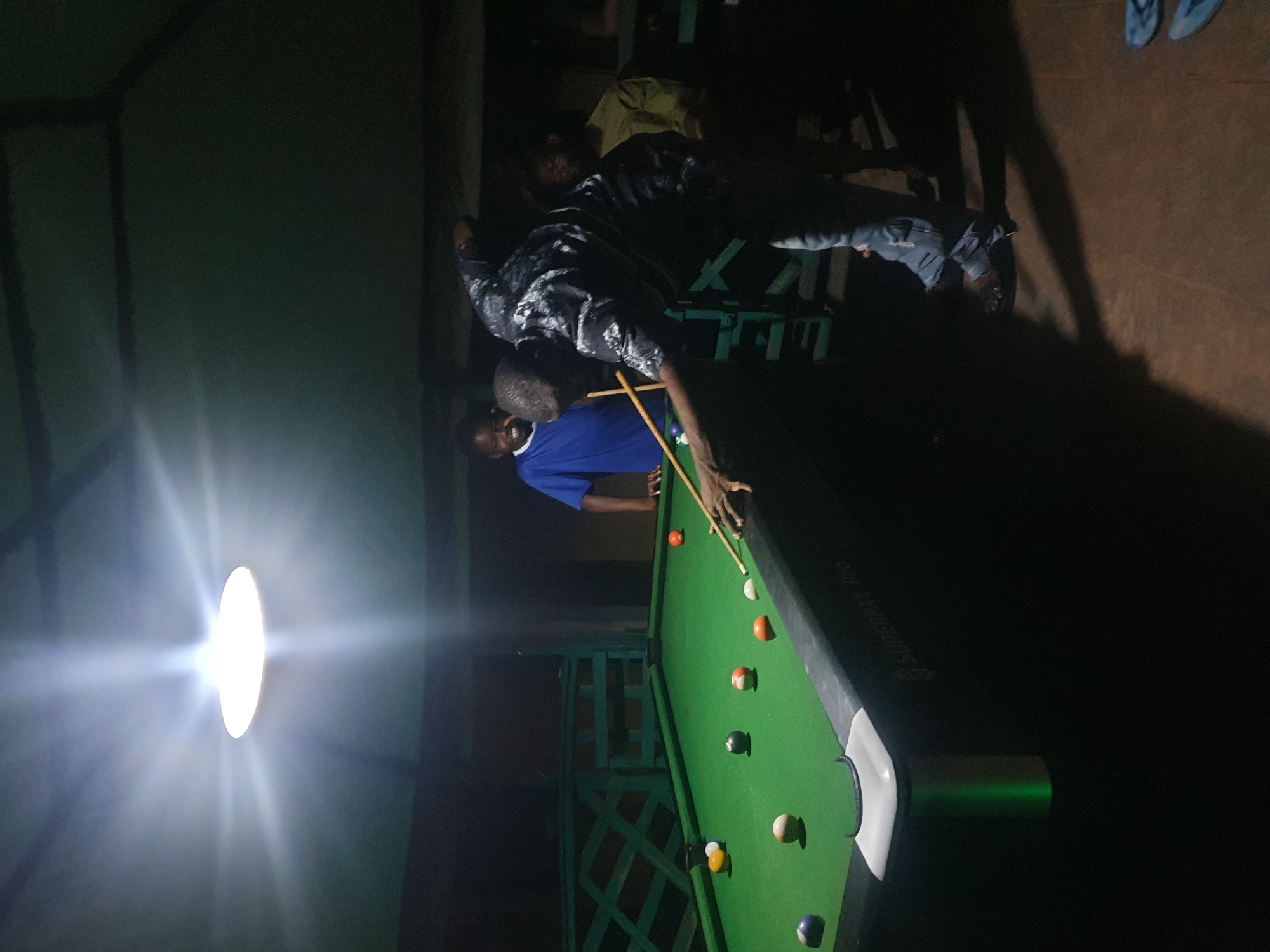 The media file shows a dimly lit indoor scene, likely a bar or recreational space, with a pool table as the central focus. The image is oriented vertically, with the pool table running diagonally across the frame.
Three individuals are partially visible. In the foreground, a person wearing a blue shirt is actively playing pool, leaning over the table with a cue stick in hand, aiming at one of the balls. Their body language suggests concentration. Behind them, another person in a dark patterned shirt is also in the process of playing pool, their stance indicating they are about to take a shot. A third person, mostly obscured by shadows and out of focus, is standing further back, observing the game or waiting for their turn.
The pool table itself is green felt with several colored balls scattered across it, including a white cue ball, striped balls, and solid colored balls. The table has a black edge and a white pocket. The lighting in the scene is uneven, with a bright, diffuse light source casting strong shadows, suggesting it might be nighttime. The background is dark and indistinct, hinting at a rustic or casual setting. There are some wooden structures and green metal supports visible in the background, possibly part of the building's architecture or furniture.
The overall mood is casual and focused on the game. The location context provided is Lokoja, Nigeria, which aligns with the somewhat informal and potentially open-air or semi-outdoor feel suggested by the background elements. The text "SUPERMAX PRO" is visible on the side of the pool table.