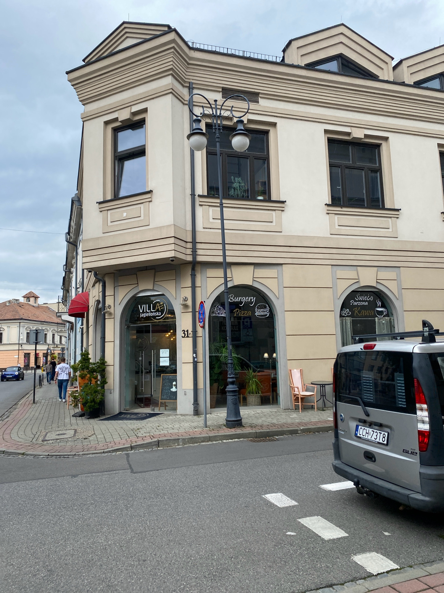 The image depicts a street corner in Nowy Sącz, Poland, featuring a multi-story building with cream-colored facade and decorative molding. The ground floor houses two establishments. The left entrance, marked "VILLA Jagiellonska", has a glass door and displays a black chalkboard menu. Next to it, an archway entrance features a sign with stylized lettering reading "Burgery Pizza". To the right, another arched entrance has a sign that says "Świeżo Parzona Kawa" (Freshly Brewed Coffee) with a coffee cup illustration. A street lamp with ornate detailing stands prominently in front of the building. A silver van with license plate "CCH 73T8" is parked on the right side of the street, partially obscuring the view of the buildings. Sidewalks are paved with cobblestones, and a road with painted crosswalks occupies the foreground. Several pedestrians are visible walking on the sidewalk in the distance, and a blue car is driving on the street. Green potted plants are placed near the entrances of the establishments. The sky is overcast.