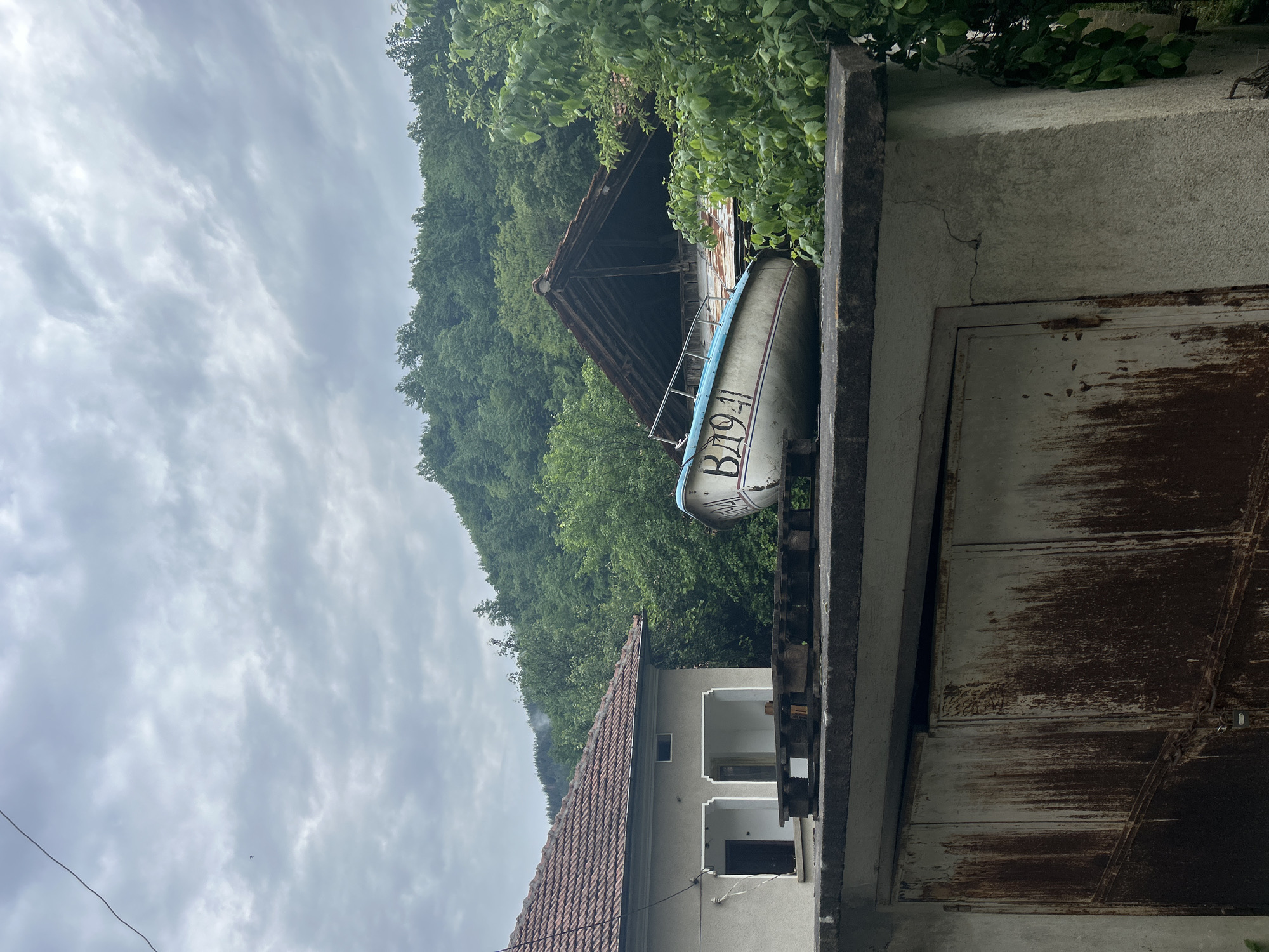 A slightly low-angle shot depicts a cluttered, perhaps rural, scene in Unknown, Unknown. A boat, labeled "B1941", is perched precariously on a concrete structure, possibly a building roof. Adjacent to the concrete structure is a rusty, metallic structure, which could be a dilapidated door or a part of a building. Further back, a house with a red-tiled roof is visible, and a wooden structure, possibly an old barn or shed, stands amongst the trees covering a hillside. The sky is overcast and grey. The lack of human presence and the disrepair of the structures suggest a neglected or abandoned setting.