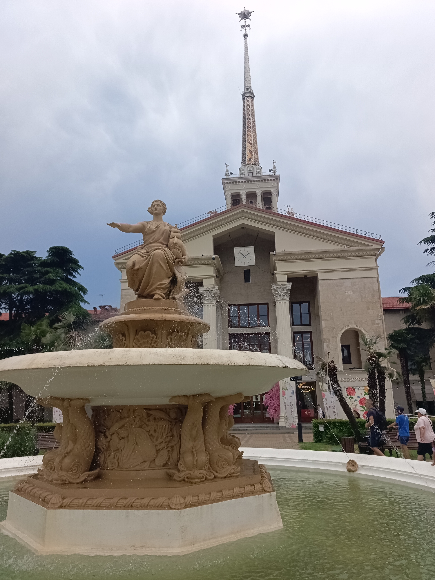 The image shows the Maritime Station in Sochi, Russia, on a cloudy day. In the foreground, a large, ornate fountain with a statue of a woman holding a child is active, with water spraying. The base of the fountain is decorated with reliefs of fish and mythological scenes. Behind the fountain, the Maritime Station building is visible, characterized by its neoclassical architecture, grand columns, and a prominent clock on its facade. A tall spire topped with a star rises from the building. Several people are in the scene, including two men and two women walking near the fountain. Palm trees and other lush greenery surround the area, suggesting a warm climate. The overall impression is of a public space, likely a tourist attraction, with a blend of art, architecture, and nature.