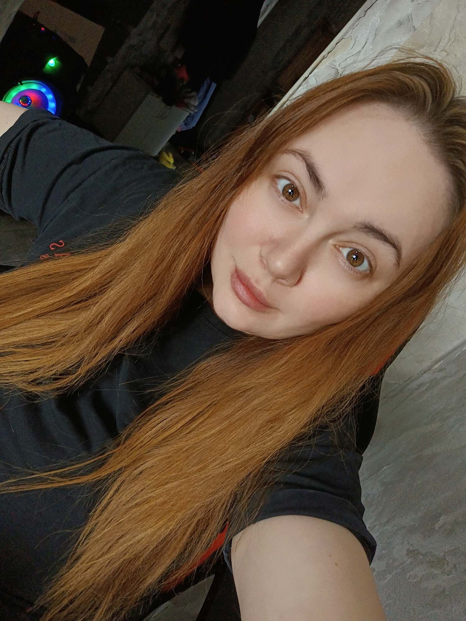 A young woman with long, reddish-brown hair and brown eyes is featured in a close-up selfie, gazing directly at the camera with a calm, neutral expression. Her head is tilted slightly, and her lips are softly parted. She has fair skin and well-defined eyebrows.
She is dressed in a black t-shirt, on which a red number "2" is clearly visible near her left shoulder, along with partial red text that appears to be another character or symbol. Her hair falls freely over her shoulders and chest.
The background indicates an indoor setting, with dim lighting suggesting it might be evening or a room with subdued illumination. To the upper left, a device with vibrant multi-colored LED lights—including red, blue, and green—is visible, resembling a speaker or a gaming accessory. The wall behind her has a textured, possibly grey or beige finish. The image captures a personal, relaxed moment. The location is Chaikovsky, Russia.