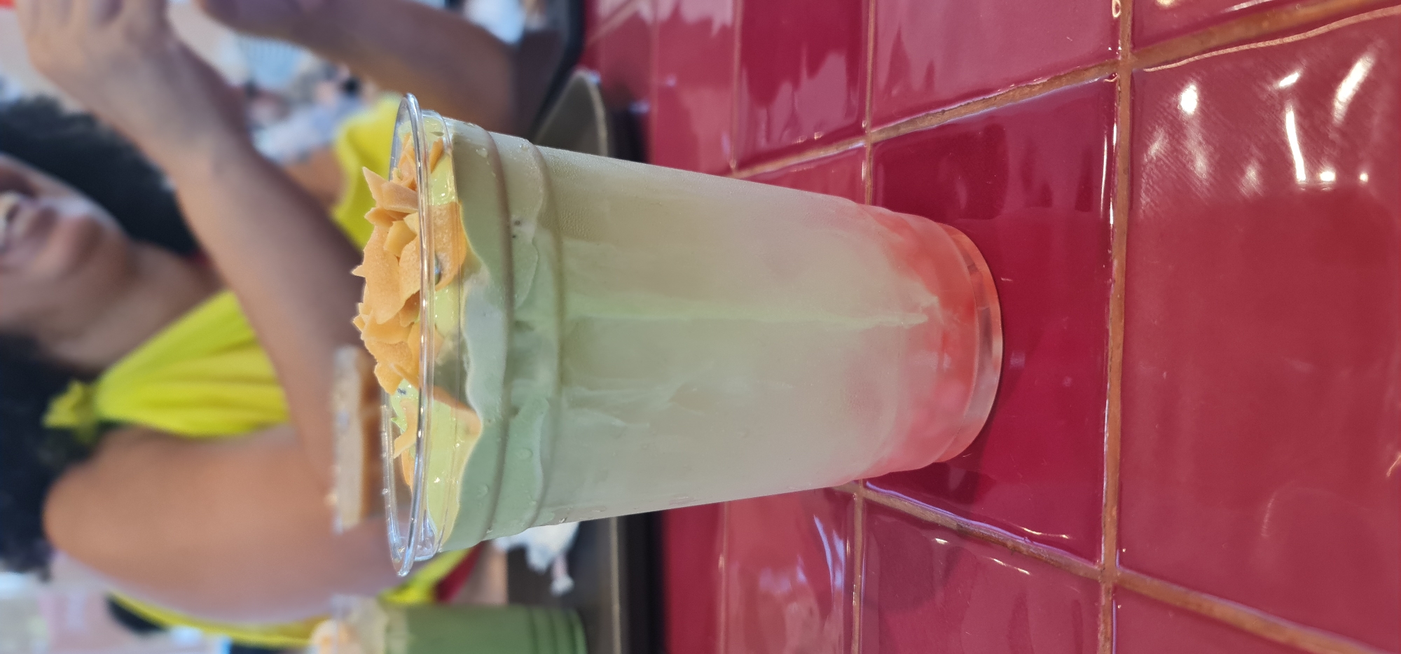 A plastic cup containing a layered beverage, possibly a smoothie or layered drink, with an orange topping is positioned on a black tray. The drink appears to have a reddish layer at the bottom and a lighter green or white layer above. The background features a wall with red, square ceramic tiles. Part of a person wearing a yellow shirt can be seen to the left of the cup. Another drink can be seen on the bottom left. The drink is placed on a table in Đà Nẵng, Vietnam.