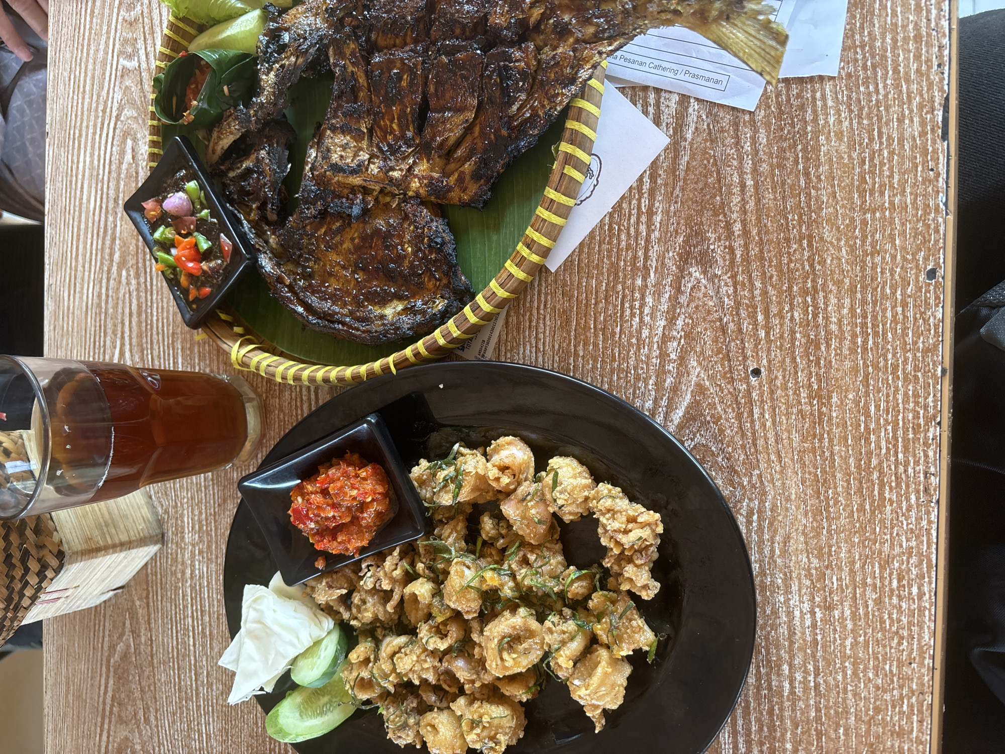 Two plates of food and a glass of iced tea are on a wooden table. One plate is black and contains fried calamari with sliced cucumbers and shredded cabbage. A small black dish holds a red chili paste. The other plate, a woven bamboo tray lined with a banana leaf, holds a whole grilled fish, a small dish of sambal, and cucumber slices. The bamboo tray has a yellow striped rim. A glass of iced tea is visible in the background, as is a woven basket. The table is made of light-colored wood with a distressed finish. The setting appears to be a restaurant, likely in West Jakarta, Indonesia, given the presence of sambal and the style of the food.