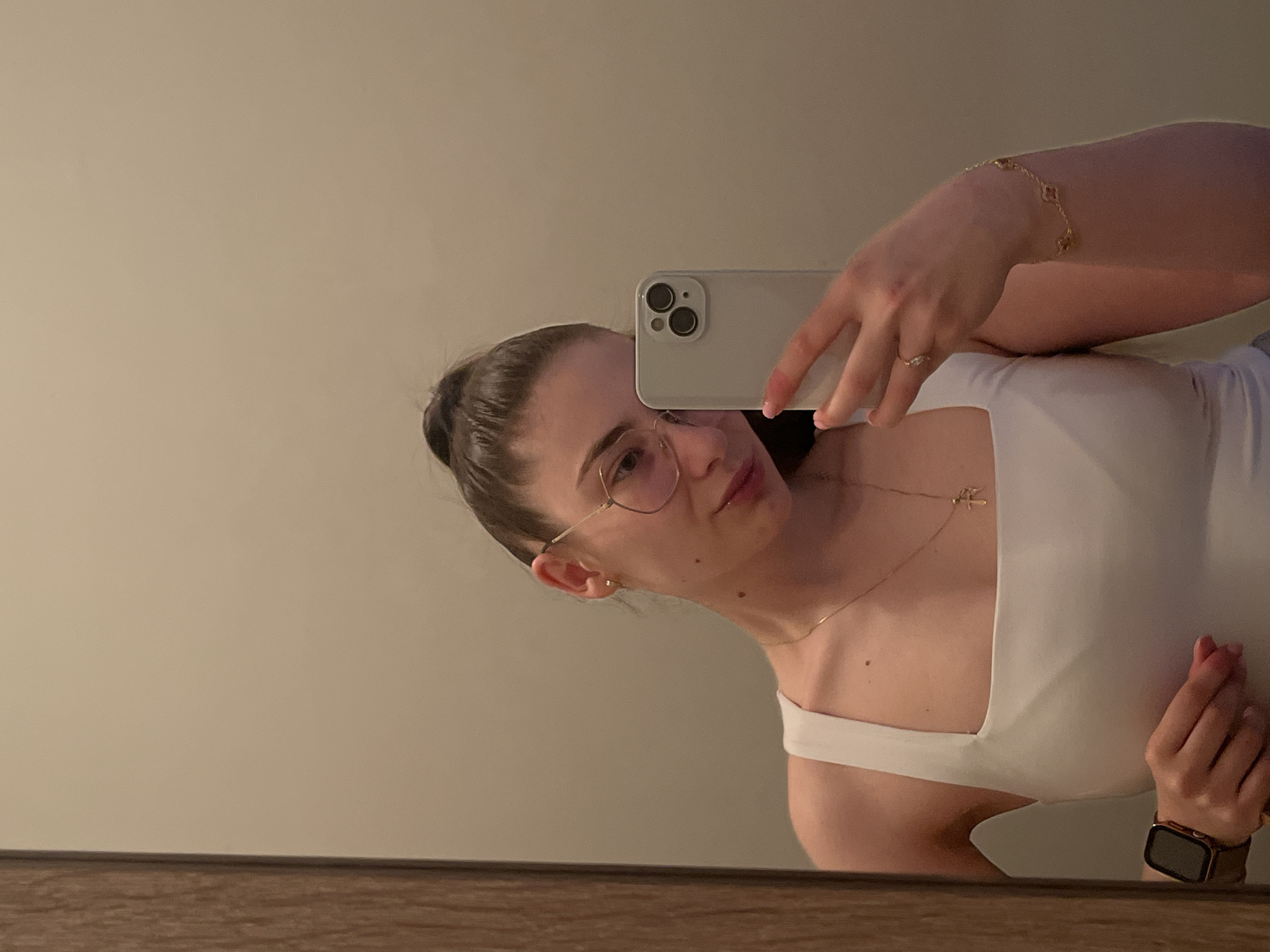 A young woman is captured in a mirror selfie, showing her reflection from the chest up. She has dark hair pulled back into a bun and wears clear-framed, possibly gold-rimmed, glasses. Her head is tilted, and she looks towards the light-colored smartphone held in her right hand, which obscures part of her face. The phone features a dual-camera setup.
She is wearing a white top, possibly a tank top or bodysuit, and accessorized with several delicate pieces of jewelry. A thin gold chain necklace with a small, unidentifiable pendant hangs around her neck. On her right wrist, she wears a gold bracelet with multiple clover-shaped charms, and a subtle ring is visible on her right ring finger. Her left wrist is adorned with a smart watch featuring a dark strap. Her fingernails have a light, natural-toned polish.
The background is a plain, light-colored wall or mirror surface, suggesting an indoor setting. A wooden surface runs across the bottom of the frame, serving as a ledge or part of the furniture. The lighting is soft and even, indicating an indoor environment, likely not under direct sunlight. The image conveys a casual, personal moment. No visible text is present.