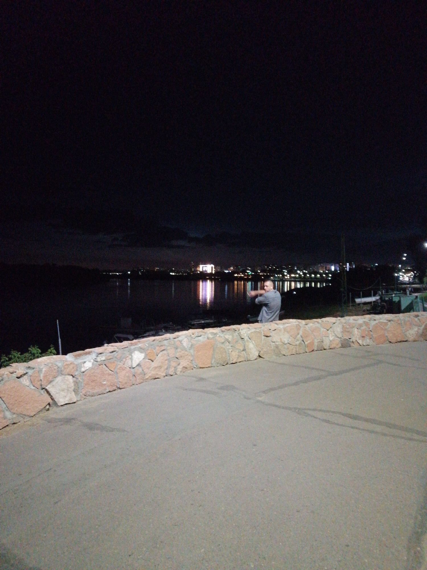 The image depicts a nighttime scene near the Irtysh River in Pavlodar, Kazakhstan. A man, wearing a grey checkered shirt, stands on a paved pathway overlooking the dark water. The city skyline is visible in the distance, illuminated by numerous lights reflecting on the river's surface. A stone wall borders the pathway in the foreground.