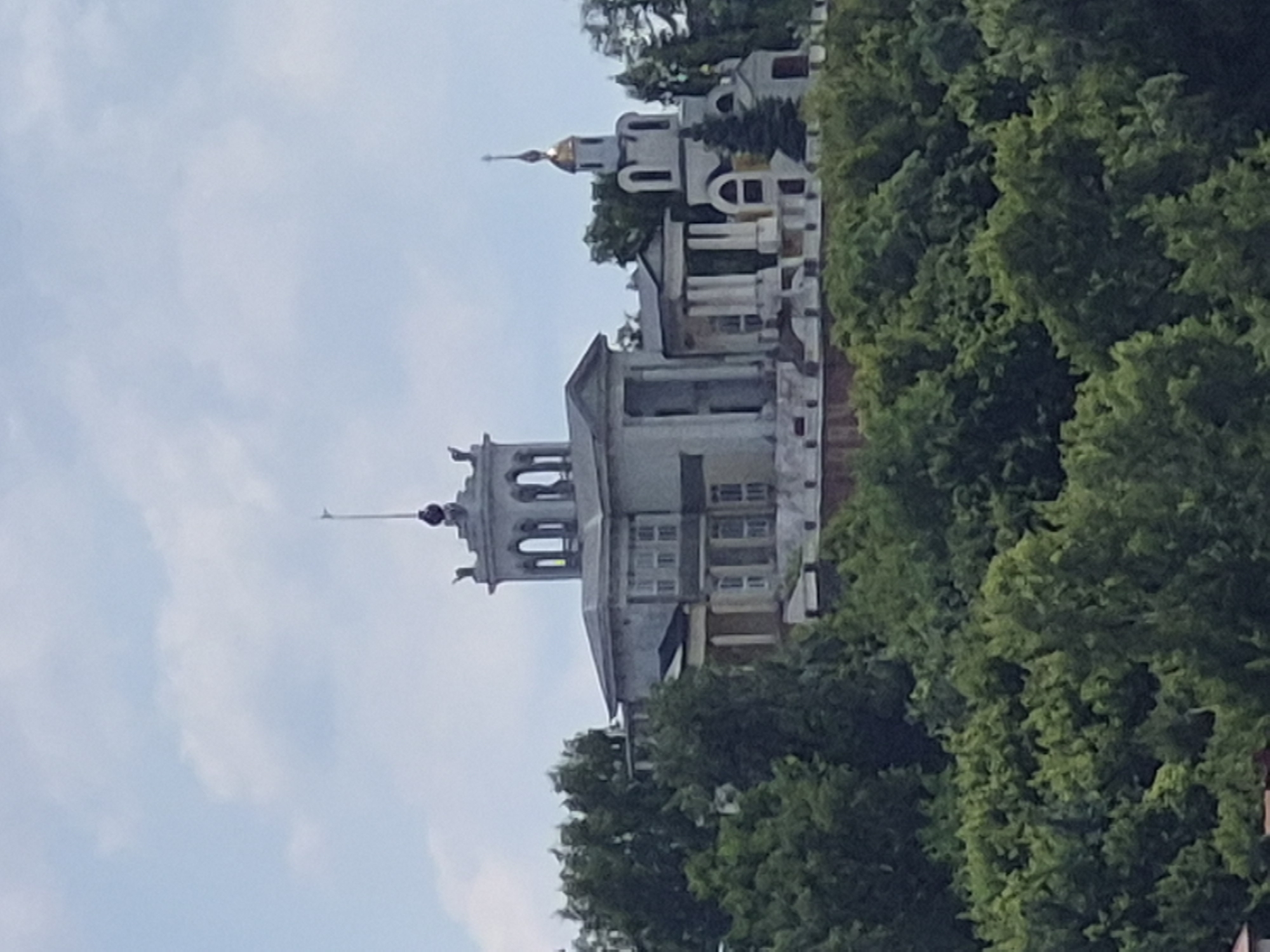 This is a medium shot, exterior photograph of the Volzhsky District Military Court in Samara, Russia, situated on a hillside overlooking a dense canopy of green trees. The architecture of the building is a mix of classical and Soviet-era styles, with a prominent central rotunda-like structure featuring arched windows and a flagpole. To the right of the main building, a smaller structure with a golden dome, characteristic of a Russian Orthodox church, is visible. The sky above is partly cloudy with a pale blue hue. The image is slightly out of focus and has a grainy texture. There are no people or visible actions depicted in the scene. The overall impression is of a historic building nestled within a lush, natural environment.