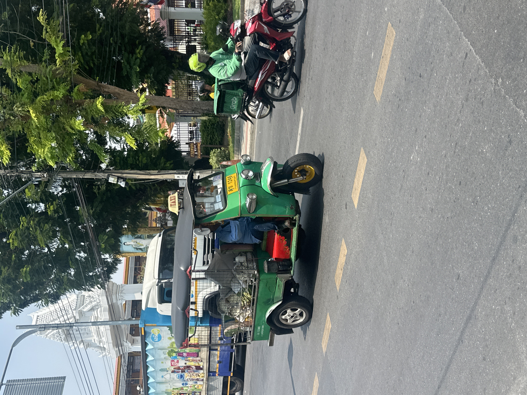 The image shows a street scene in Bangkok, Thailand, with a green tuk-tuk prominently displayed in the center. The tuk-tuk, a three-wheeled motorized vehicle, is filled with various items in its rear cargo area. A motorcycle with two riders, one wearing a green jacket and helmet and carrying a "Grab" delivery bag, is next to the tuk-tuk. The street is marked with yellow lane lines. In the background, there is a building with white architectural details, a tree, and utility poles with wires. A van and an adjacent storefront are visible on the left side of the image, displaying items for sale.
