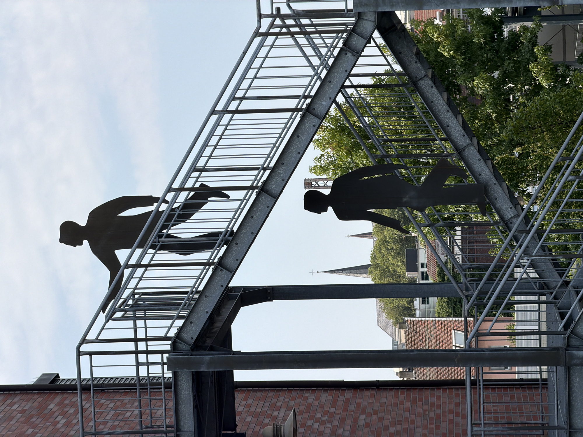 Two black silhouette figures of men are depicted on a metal fire escape stairwell. The upper figure is walking upwards, with their right leg extended. The lower figure is walking downwards, with their left leg extended. The fire escape is attached to a brick building on the left side of the image. In the background, there are buildings with tiled roofs, green trees, and two church spires. The sky is cloudy and light blue. This fire escape is located at the "Archäologisches Museum Bochum" in Bochum, Germany.
