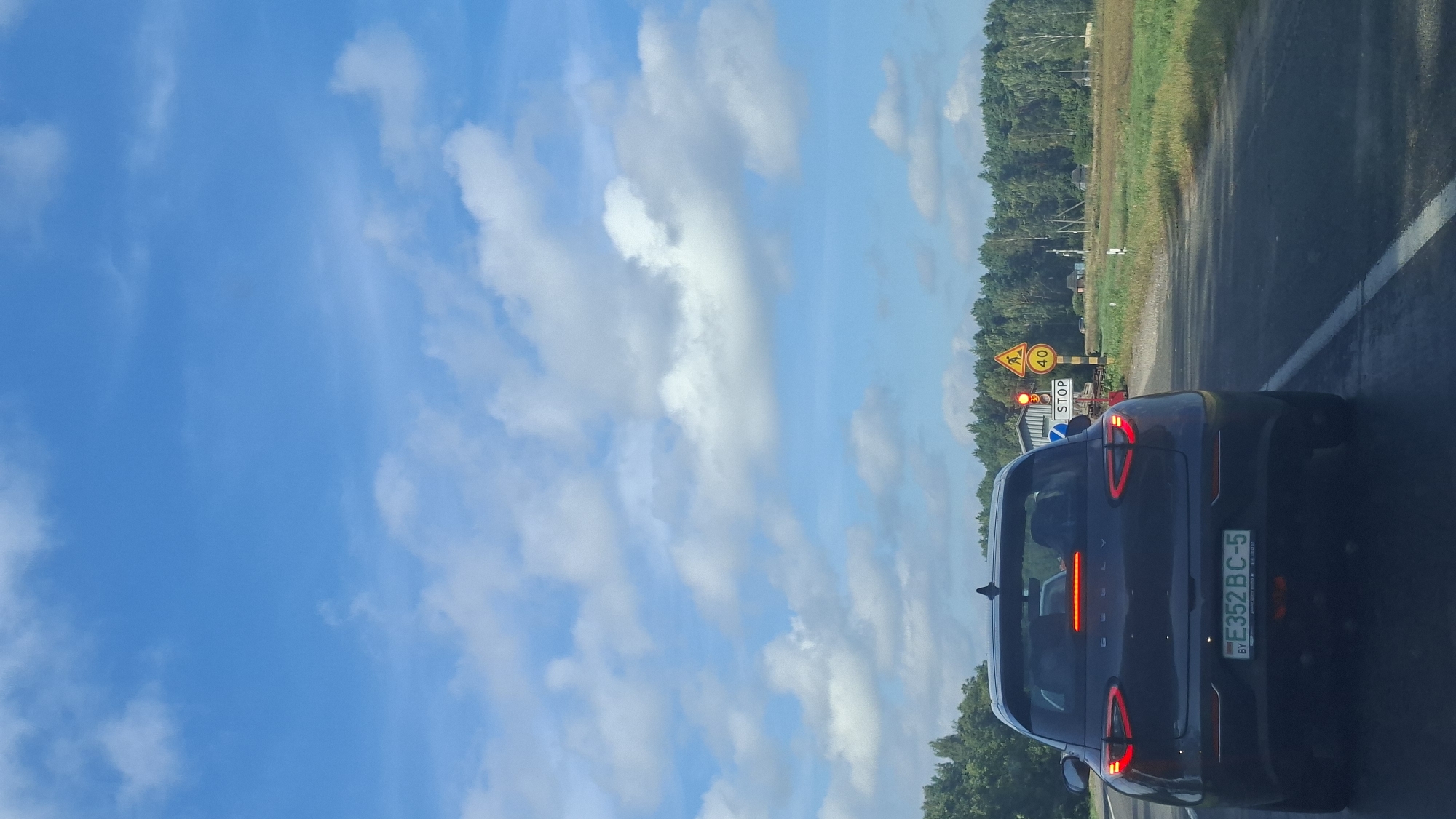 The image captures a view from the rear of a dark-colored Geely electric vehicle driving on a paved road. The car's brake lights are illuminated. The license plate is visible and reads "BY E352 BC-5". The road is bordered by a grassy shoulder and a dense line of green trees in the background. In the distance, there are traffic signals including a red traffic light, a sign indicating "STOP", a triangular sign with a pedestrian symbol, and a circular sign displaying "40". The sky above is a bright blue with scattered white clouds. The scene appears to be in a rural or semi-rural area, consistent with the location of Асіпавіцкі сельскі Савет, Belarus.