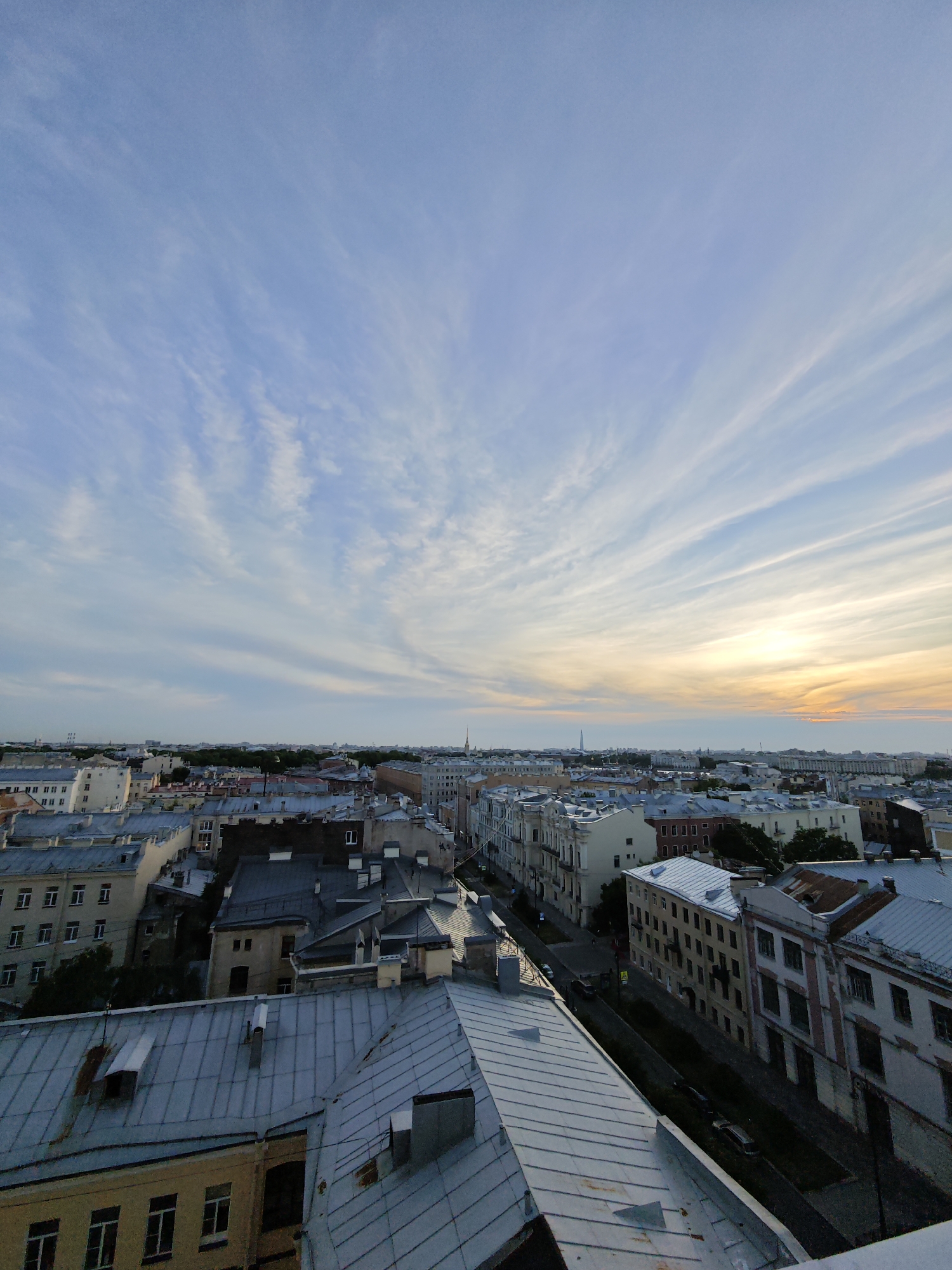 An aerial, high-angle, vertical shot showcases a sprawling cityscape of Saint Petersburg, Russia, bathed in the soft, warm glow of a setting sun. The sky is a canvas of pale blue, streaked with wispy, cirrus clouds that stretch horizontally across the frame, catching the last rays of sunlight in hues of orange and yellow. Below, a dense cluster of buildings with distinct St. Petersburg architecture fills the midground and foreground. The buildings are primarily a muted palette of grays, beige, and white, with many featuring steeply pitched metal roofs, some appearing aged and weathered. The rooftops are a dominant feature, creating a complex, textured landscape of varying shapes and slopes. Streets are visible between the buildings, with the presence of parked cars and what appear to be street lamps. The overall impression is one of a historic urban environment at the end of the day, with a sense of calm and vastness conveyed by the expansive sky and the layered cityscape.