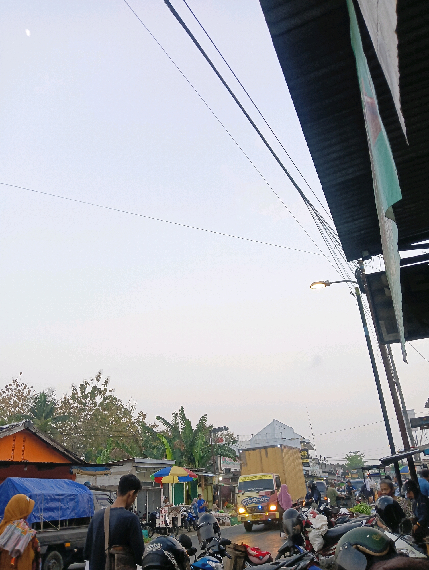 The image depicts a street scene in Purwareja, Indonesia, likely a market area, with a mix of vehicles, people, and market stalls under an overcast sky. A yellow truck with a cargo container is prominent in the mid-ground, its headlights on. Several motorcycles are parked on the right side of the street. People are visible standing and walking around market stalls, some appearing to be vendors and others customers. In the foreground, the back of a person wearing a dark shirt and a bag is visible. To the left, a truck covered with a blue tarp is parked. Overhead, power lines crisscross the sky, and a street light is illuminated. Buildings with orange and grey roofs line the street, interspersed with lush green trees and banana plants. The overall atmosphere suggests a busy, active commercial zone during daylight hours.
