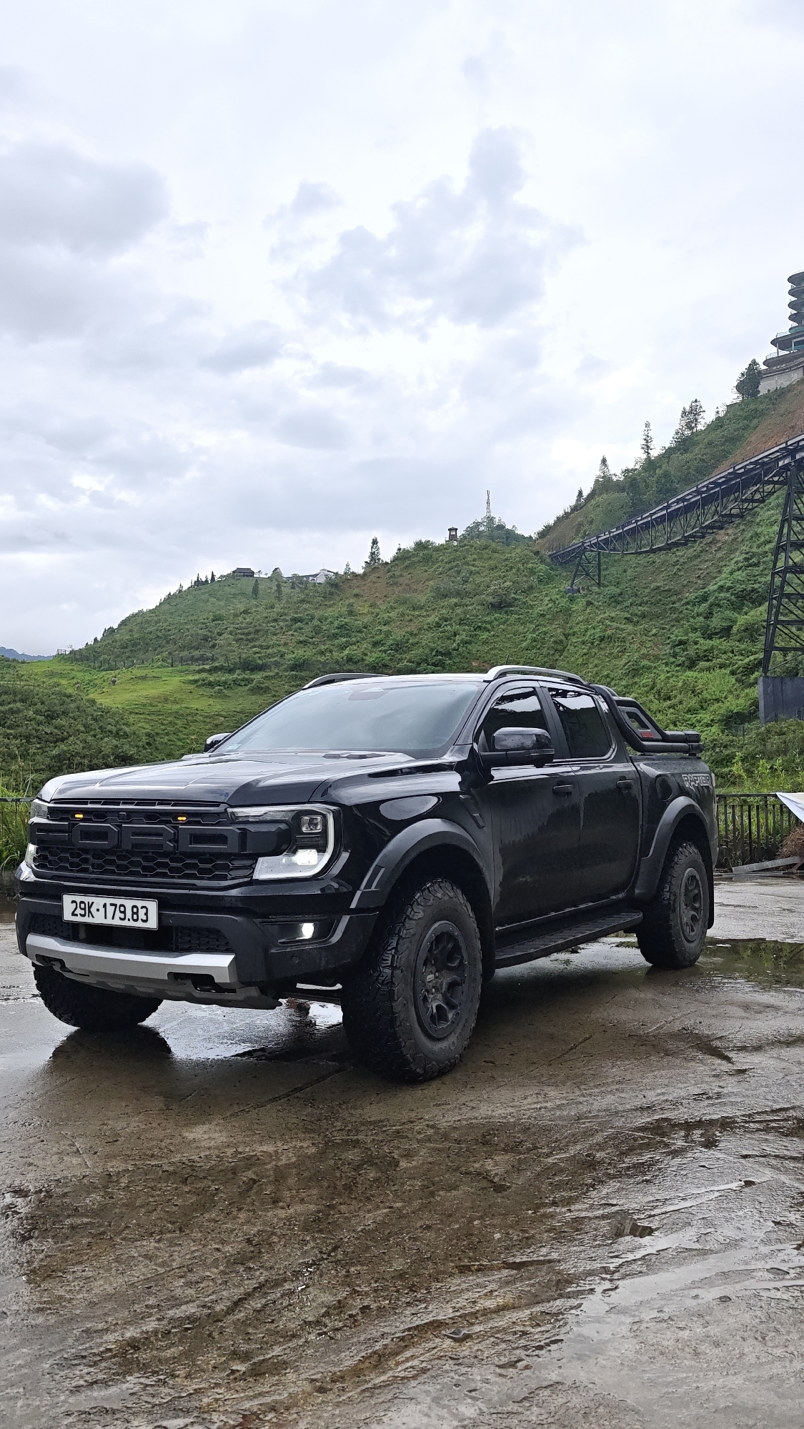 The image shows a black Ford Ranger Raptor pickup truck parked on a wet concrete surface. The truck has a license plate that reads "29K-179.83". The background features a green hillside with a metal structure resembling a cable car system. In the distance, there is a building or structure on the hilltop. The sky is overcast, suggesting cloudy weather. The scene is likely located in a mountainous or hilly area. Based on the visible elements, the location is consistent with the scenery found in Cat Cat Village, Vietnam, which is known for its mountainous terrain and cable car systems.