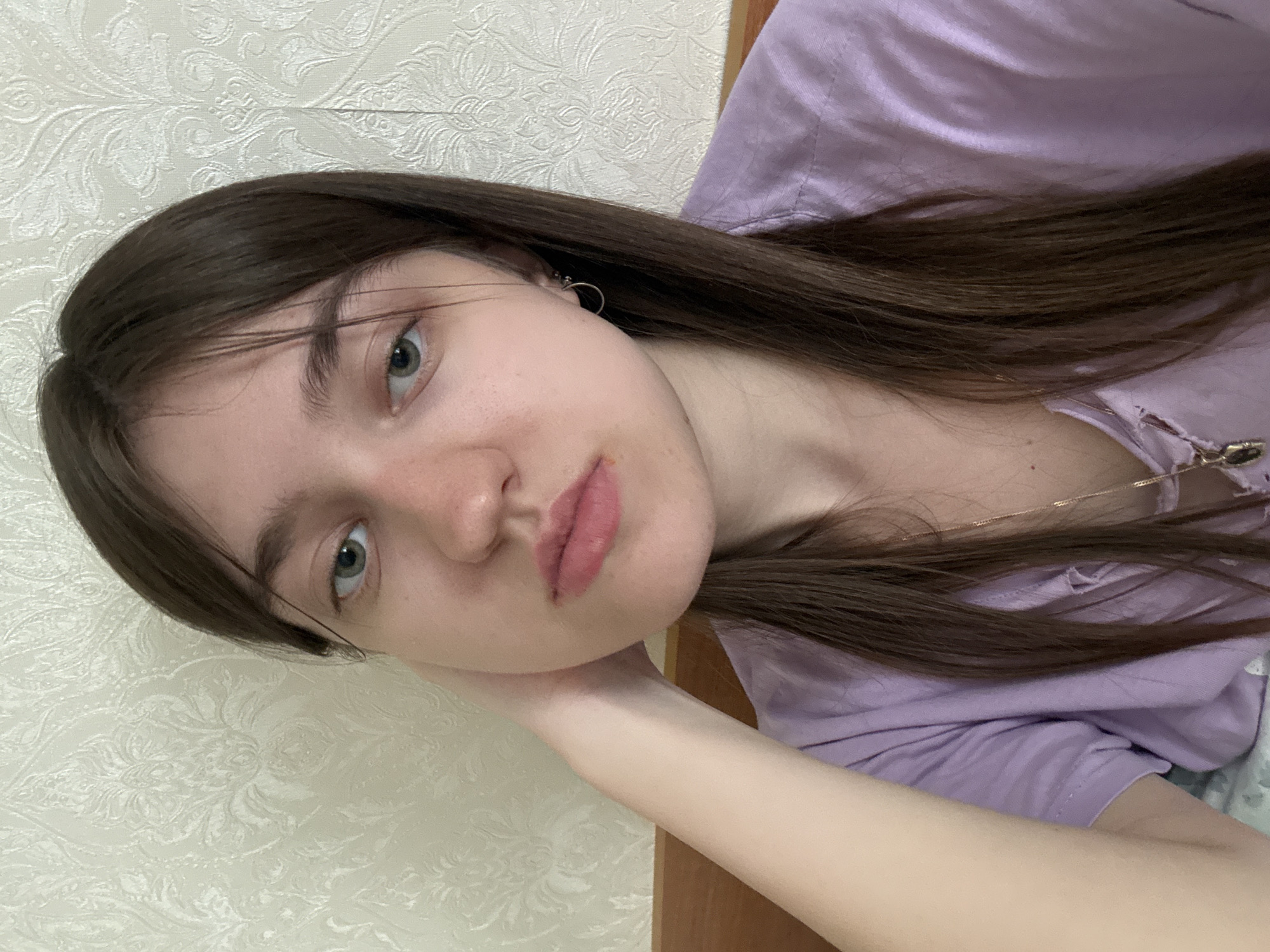 A close-up, indoor, portrait shot depicts a young woman with long, straight, dark brown hair. She has fair skin, light-colored eyes, and is wearing a light purple shirt. A thin, gold necklace is visible around her neck. She is resting her head on her hand. The background consists of a textured, light-colored wall. The location is not specified.
