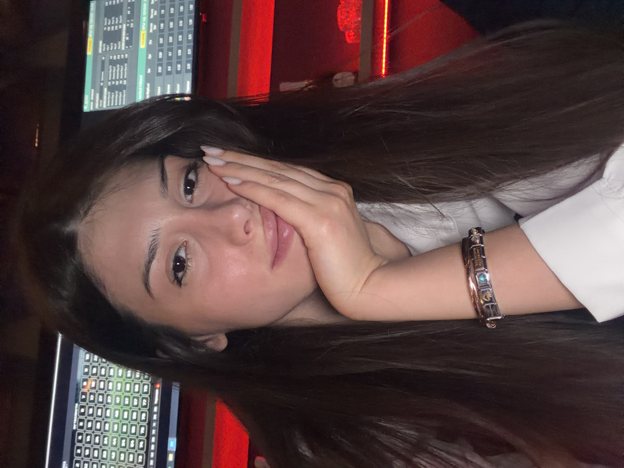 A young woman with long, dark hair rests her cheek on her hand, her eyes looking towards the camera. She wears a white collared shirt and a silver bracelet with blue and green charms. In the background, two screens display data, and red lighting illuminates the shelves behind her, which hold bottles and glasses. The scene suggests an indoor setting, possibly a bar or a place with electronic displays.