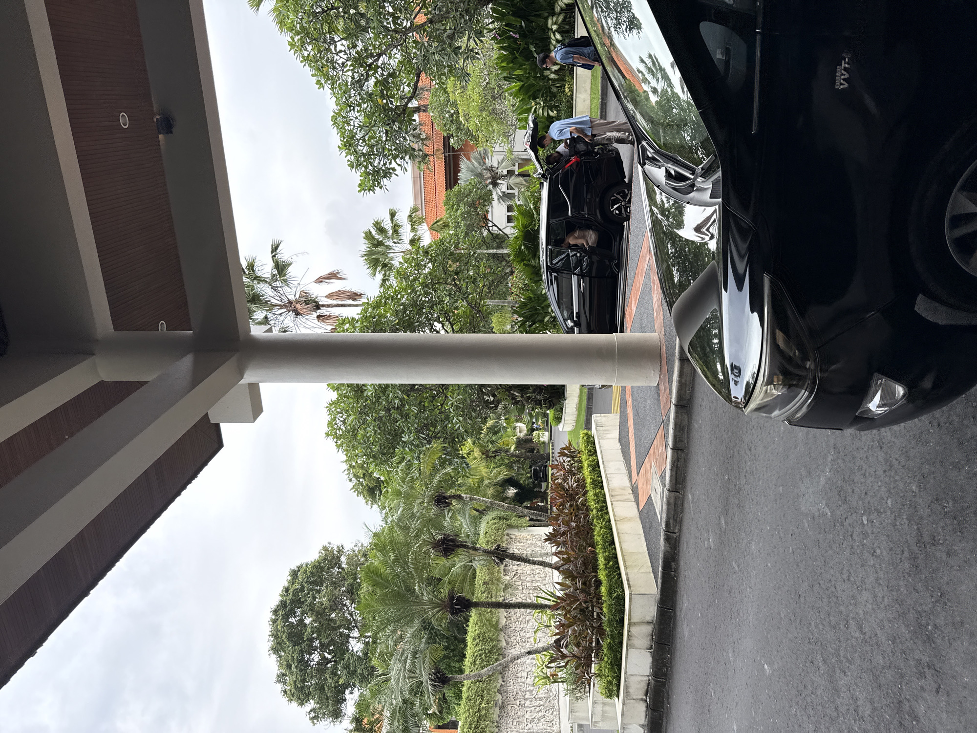 The scene is outdoors in Nusa Dua, Indonesia, showcasing a hotel or resort driveway. A black vehicle is parked on the right side of the frame, taking up a significant portion. A second, similar vehicle is parked nearby with two people standing adjacent, one appearing to assist someone sitting inside. Tropical greenery and palm trees surround the area, adding to the resort-like environment. Part of a building structure, likely the hotel entrance, is visible on the left with its architectural details.