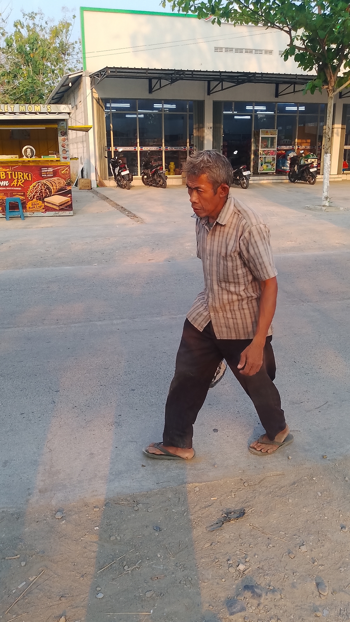 The image shows an elderly East Asian man walking on a paved surface in what appears to be an outdoor commercial area. He is wearing a short-sleeved, vertically striped button-down shirt and dark pants. His footwear consists of dark-colored flip-flops. The man's facial expression is neutral as he walks towards the right side of the frame.
In the background, a building with large glass windows and a green roof trim is visible. Several motorcycles are parked in front of the building. To the left of the building, there is a food stall with signage, including text in Indonesian, such as "MOM'S" and "BT TURKI AR". A blue stool is placed near the food stall. The ground is a mix of paved areas and dirt. The lighting suggests it is daytime, possibly late afternoon, with long shadows cast on the ground. The environment appears to be in Karanglangu, Indonesia, based on the provided context, with elements of a commercial street scene.
