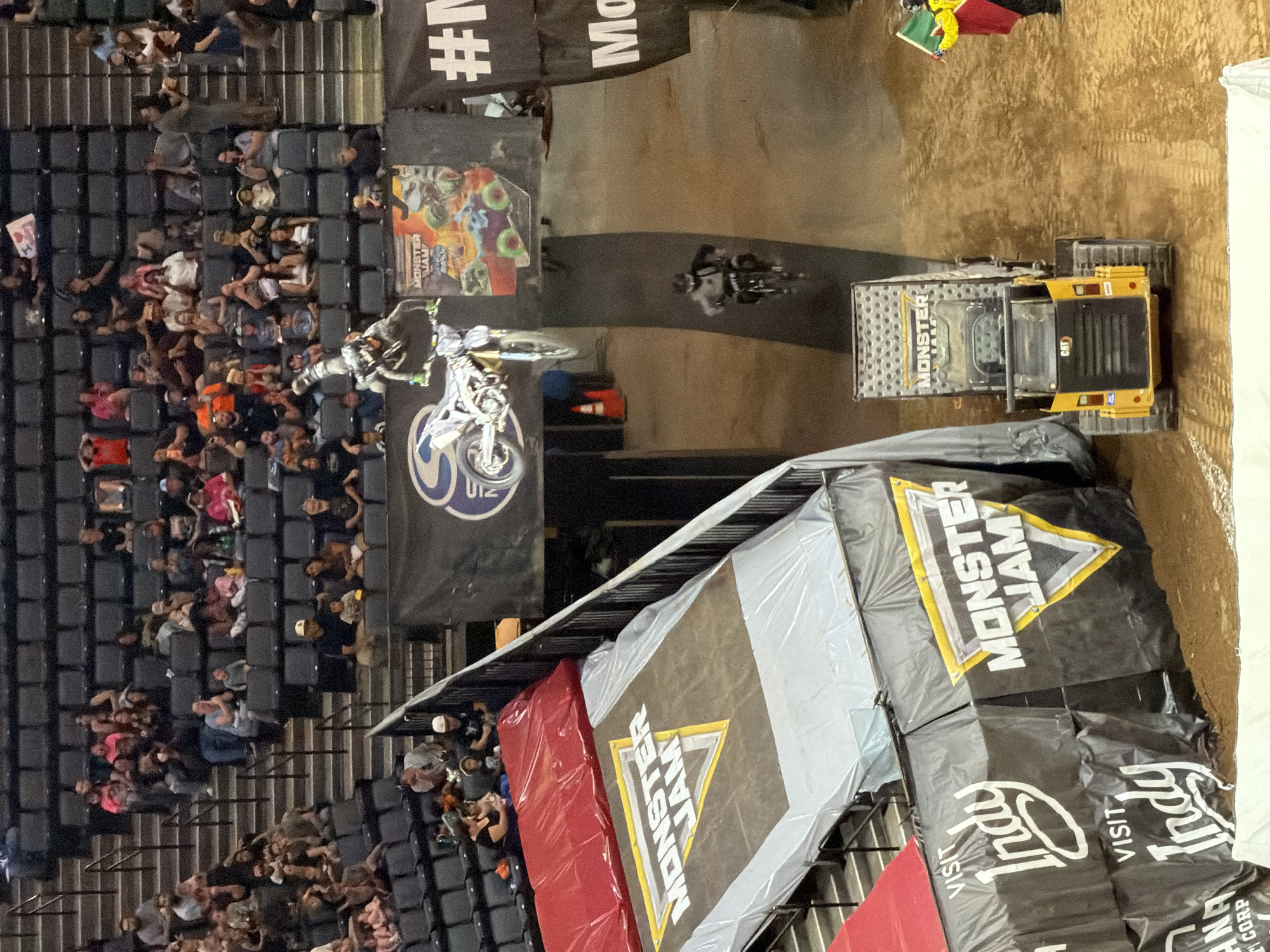 The image depicts a motocross event, likely Monster Jam, taking place indoors in Sacramento, California, USA. The scene is dominated by a dirt track and ramps, with a motocross rider in mid-air performing a jump on their bike. In the background, spectators are seated in tiered stadium seating, observing the event. Several banners and signage, including "MONSTER JAM" and "VISIT Indy," are visible around the venue, indicating sponsorship and location details. A yellow construction vehicle, possibly a skid steer loader, is present on the dirt track, and a person holding a flag stands to the right of the vehicle. The lighting suggests an artificial indoor environment.