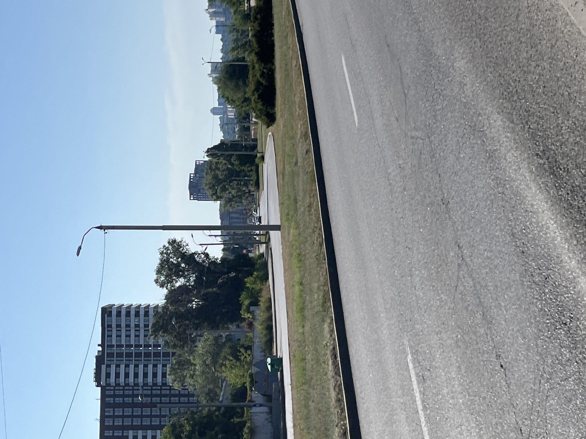 The image depicts a wide, paved road curving through an urban landscape under a clear blue sky. In the foreground, the road surface shows some wear and tear, with a white lane marker visible on the right side. To the left of the road, a strip of dry, green grass is bordered by a dark curb. Beyond the grass, a low concrete wall separates the road area from a more distant plane. On this plane, a bright green trash bin is situated near the wall, and further back, a line of trees and shrubs forms a green buffer.
Dominating the left side of the midground is a tall, modern apartment building with a dark facade and numerous windows. Several streetlights with distinct curved arms are positioned along the road. In the distance, a cityscape unfolds with more buildings of varying heights and architectural styles, some appearing to be residential towers and others possibly commercial structures. The overall impression is one of a sunny day in a developed urban area.