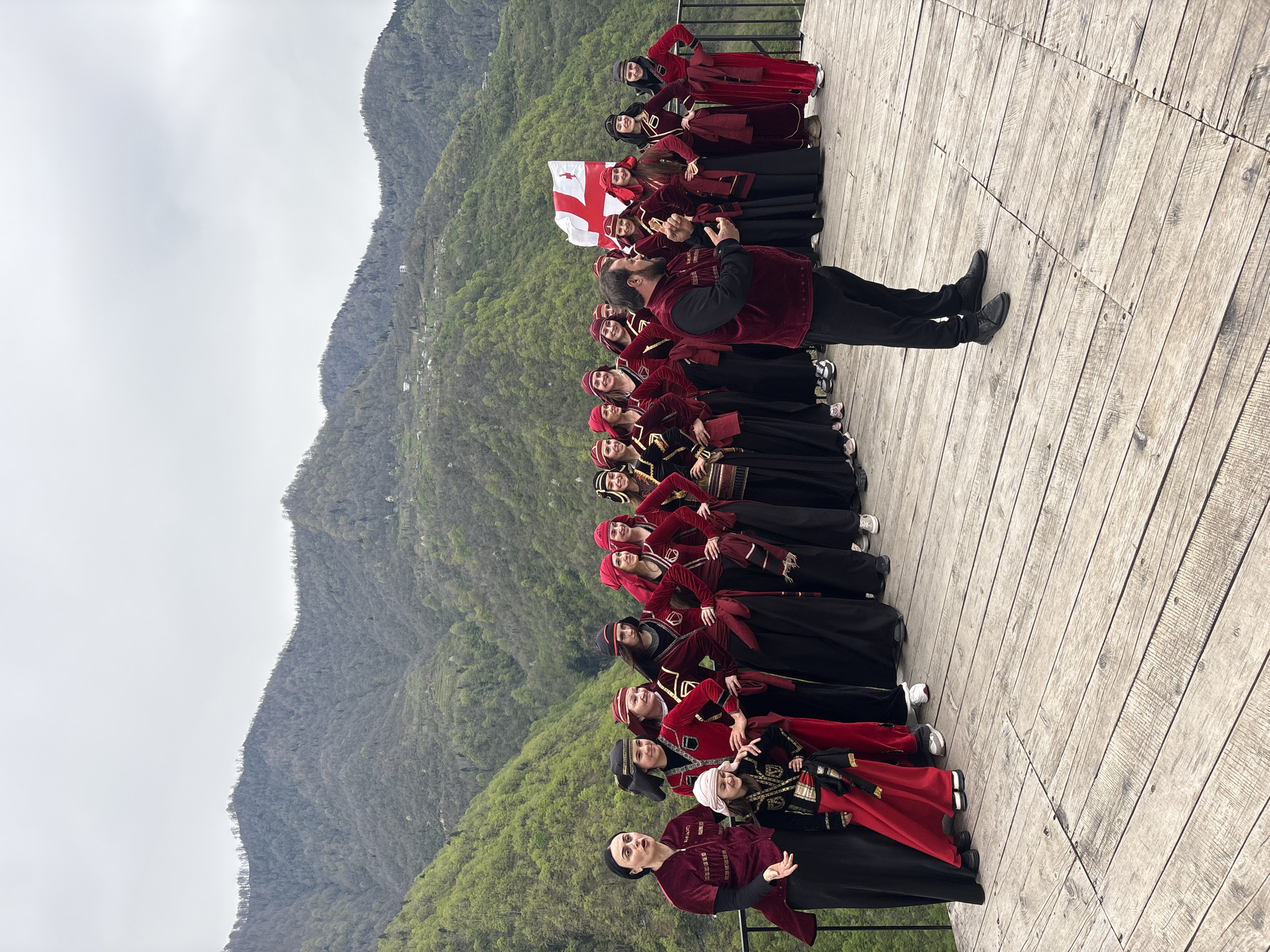 A wide-angle outdoor photograph captures a group of people posing on a wooden deck against a backdrop of mountains. The group consists of women and one man, all dressed in traditional Georgian clothing featuring red and black colors. The man appears to be leading or directing the group, gesturing with his hands. A Georgian flag is held aloft towards the back of the group. The deck is made of light-colored wooden planks and stretches across the lower portion of the frame. Behind the group, a mountain range covered in green vegetation rises towards a cloudy sky. The city of Kveda Chkhutuneti, Georgia is not visible in this image.