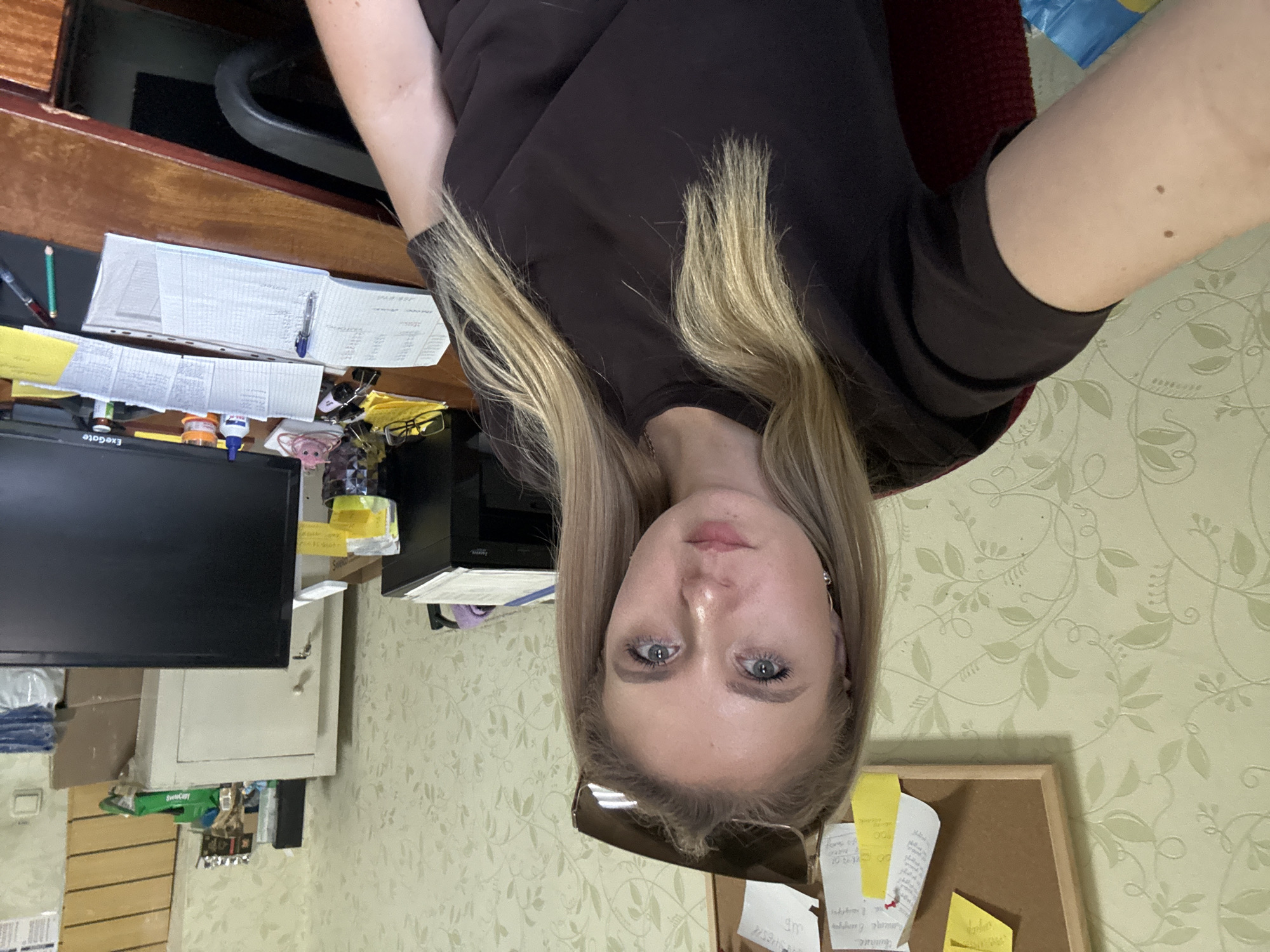 The media file is a selfie taken in an office or study setting, likely during the daytime as indicated by the ambient lighting. The main subject is a young woman with blonde hair, who appears to be in her late teens or early twenties. She is positioned in the foreground, with her face and upper torso visible. She has blue eyes, fair skin, and is wearing a dark-colored t-shirt. She is looking directly at the camera, and her expression appears neutral. Sunglasses are perched on her nose.
The background reveals a cluttered desk with various office supplies. To the left, a computer monitor is visible, along with stacks of papers and what appears to be a binder. Further to the left, part of a wooden desk and a chair are visible. To the right, there is a bulletin board with several notes and pieces of paper pinned to it. The wall behind the desk is covered in a light green patterned wallpaper. Some of the visible text on the papers includes handwritten notes and what appears to be some form of ledger or spreadsheet. One note on the bulletin board includes the text "SWEET COPY". Another visible item is a small pink trinket. The overall scene suggests a typical workspace environment.