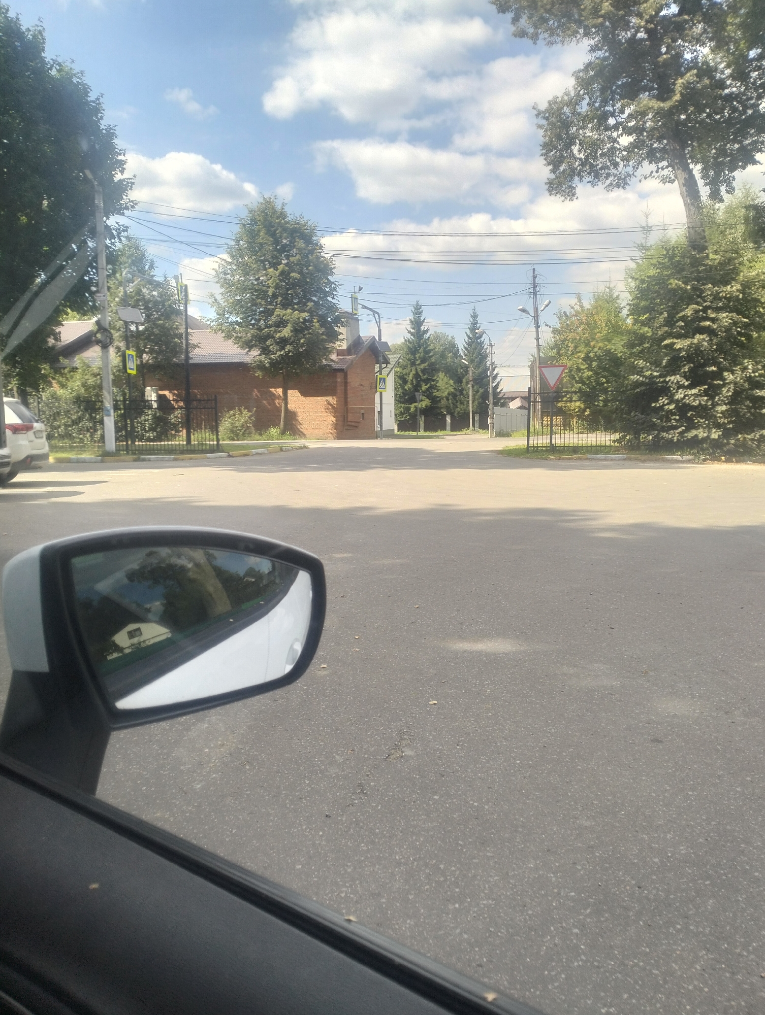 The image is a medium shot of a residential street in Osechenki, Russia, taken from inside a vehicle. The perspective is from the driver's side, with the side-view mirror prominently displayed in the lower left corner, reflecting a clear blue sky with scattered clouds and some greenery. The road in the foreground is paved asphalt. In the background, there are brick houses with brown roofs, trees, and utility poles with overhead wires. A yield sign is visible on a utility pole. To the left, a white car is parked on the side of the road. The scene is illuminated by sunlight, suggesting it is daytime.