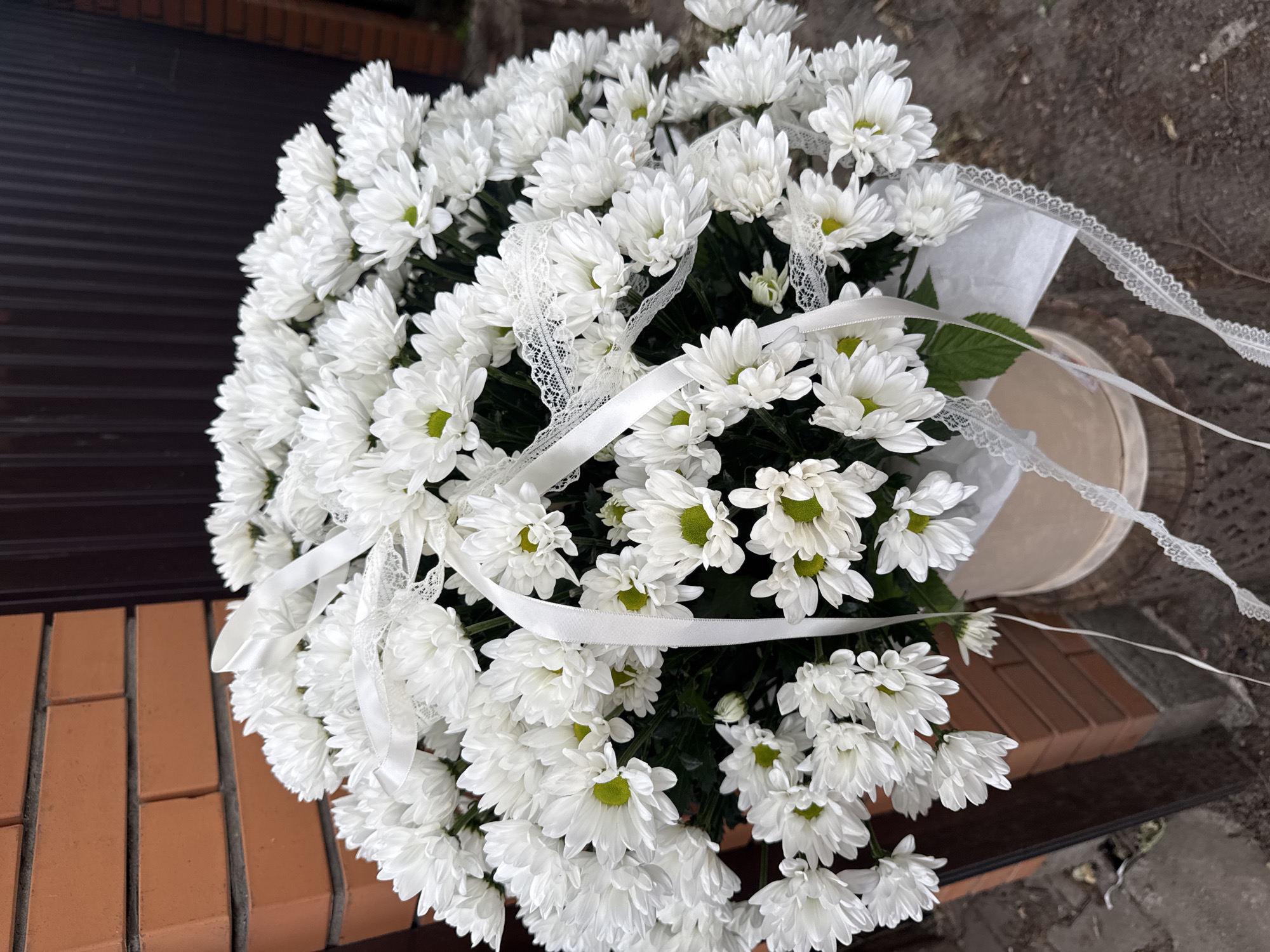 A large bouquet of white chrysanthemums is presented in a light-colored box, adorned with white satin ribbon and delicate lace. The bouquet is positioned outdoors, with a brick wall and a dark fence visible in the background. The ground appears to be paved or covered with gravel. The image does not contain any people or discernible clues to pinpoint its specific location within the city of Almaty, Kazakhstan.