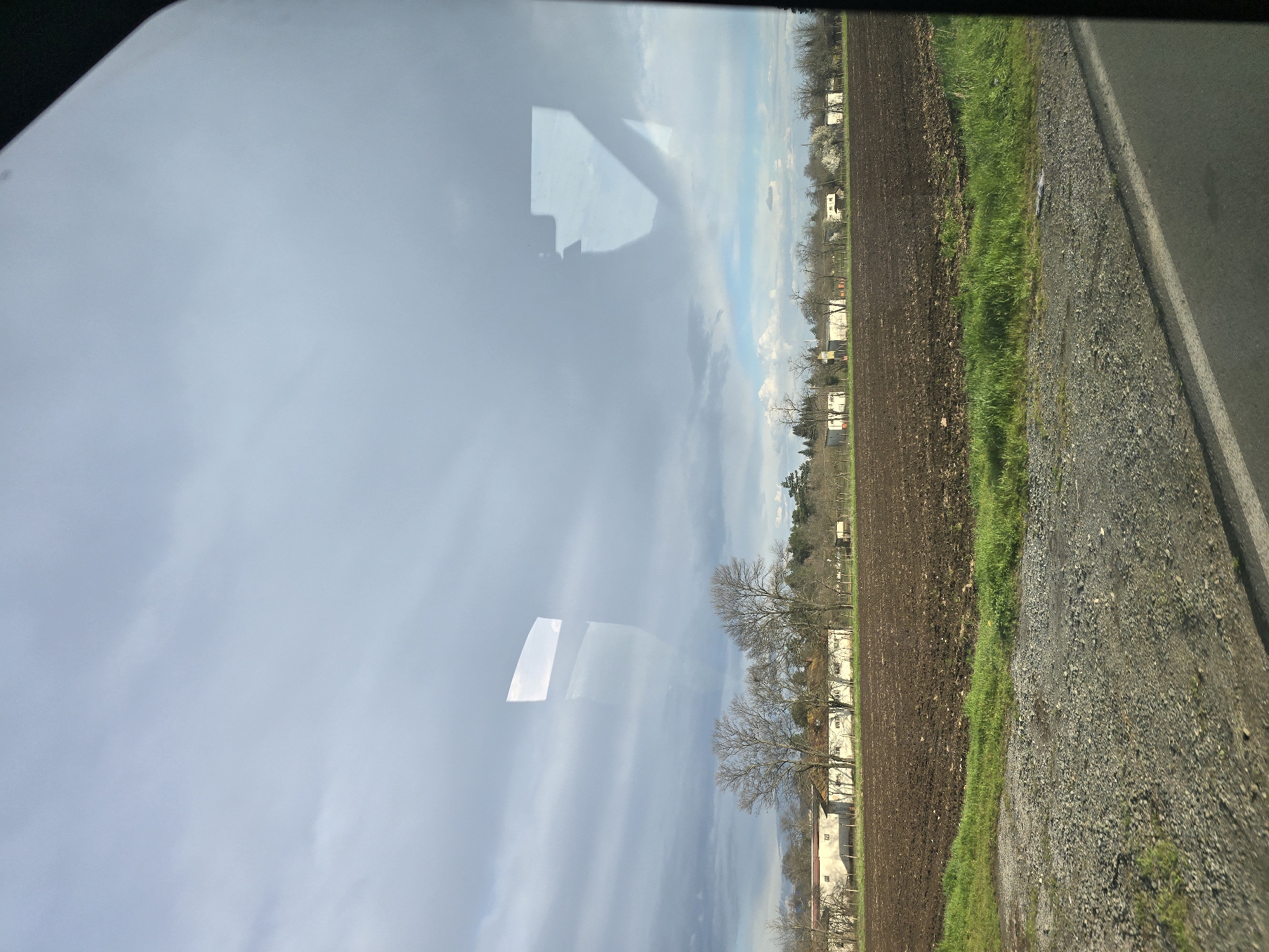 The image shows a rural scene viewed from inside a vehicle, possibly a car. The location is likely in or near Kabile, Bulgaria, given the provided context. The sky is overcast with scattered clouds. Reflections are visible on the window of the vehicle. The ground consists of a freshly plowed field, transitioning to a grassy verge, followed by a gravel road and then a paved road. Buildings with light-colored walls are situated near the edge of the field, with bare trees scattered among them. A gray and white bird is lying in the grass.