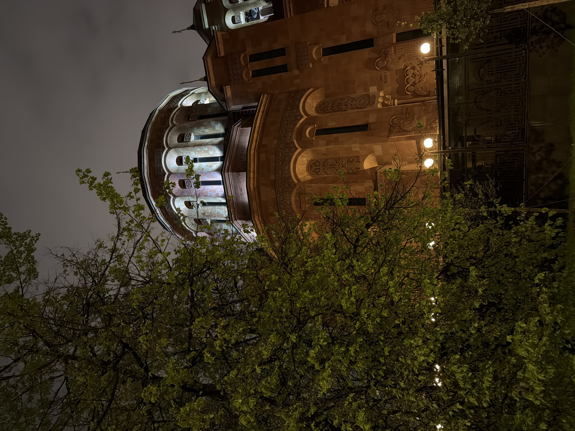 The image shows the Armenian Cathedral Complex in Moscow, Russia, at night. The cathedral's illuminated exterior is visible above trees, revealing its distinct architectural features: a rotunda-like structure and a taller, adjacent building with decorative carvings. The scene includes illuminated windows and ornate details on the facade, typical of Armenian church architecture. Streetlights can be seen among the trees, adding to the nighttime illumination. The sky appears overcast.