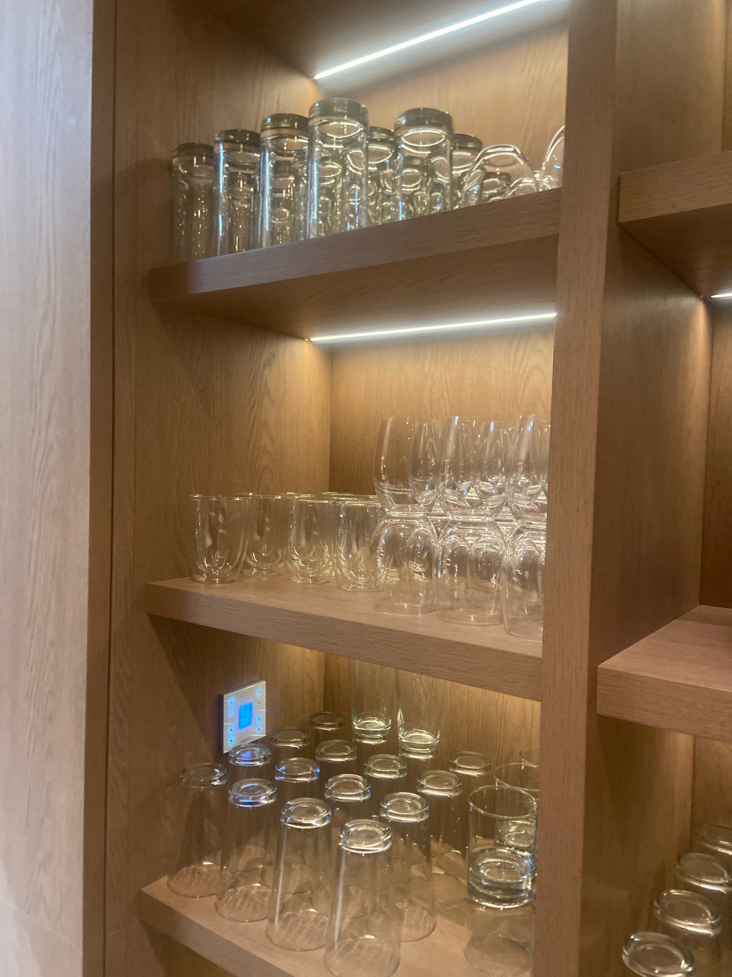 The image displays a well-lit, modern wooden shelving unit filled with various types of glassware. The shelves are organized with neatly stacked drinking glasses and wine glasses. Some shelves have LED strip lighting installed at the front edge, illuminating the glassware. On the second shelf from the bottom, a small digital control panel with a blue display is visible on the left side. The overall setting appears to be an interior space, possibly a kitchen, bar area, or a dining room. The image does not provide any explicit visual cues that would allow for its specific location in Ngaglik, Indonesia.