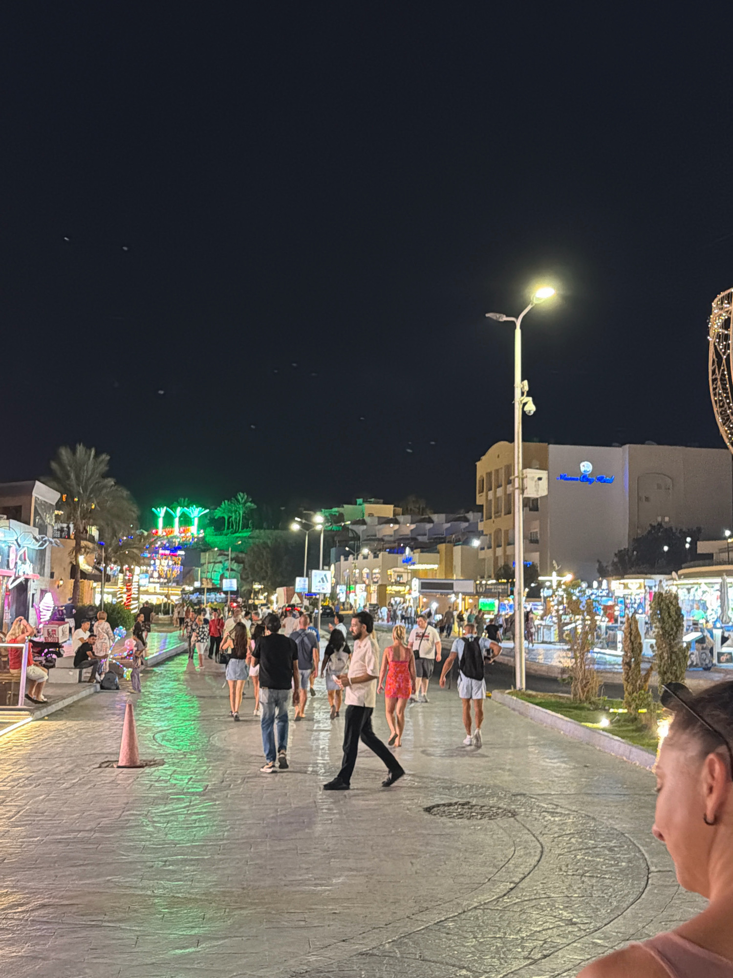 This is a nighttime, outdoor, eye-level, medium-wide shot of a busy pedestrian street in Sharm El Sheikh, Egypt. The street is paved with light-colored, textured stones, and illuminated by streetlights and colorful neon signs. People are walking in both directions on the street, some individually and some in small groups. Several buildings are visible on either side of the street, some of which appear to be shops or restaurants with their lights on. In the background, there are palm trees and more buildings, some with neon lights. The overall atmosphere is lively and festive, suggesting a popular tourist area.