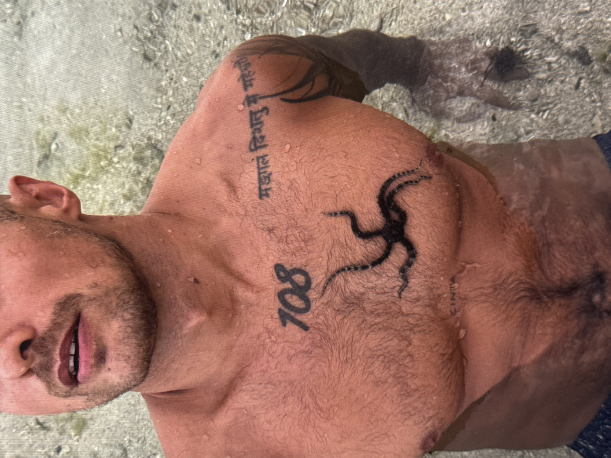 This is a close-up, overhead shot of a person lying in shallow, clear water. The water is calm, with a sandy, pebbly bottom visible beneath the surface. Sunlight is refracting through the water, creating dappled patterns.
The subject is a person with tanned skin, likely male, with chest hair and water droplets on their skin. They have several tattoos: a vertical line of text in a non-Latin script on the upper left chest, an octopus-like design on the center chest, and the numbers "708" in large font below the octopus. Faint text, possibly "FINITA," is visible on the lower right chest. The person's head is partially visible on the left side of the frame, with an open mouth and a beard.
The activity appears to be relaxation or leisure in a natural water setting, possibly a beach or a shallow sea. The overall mood is serene and peaceful. The location context provided is Kuta Selatan, Indonesia, which suggests a tropical environment. The time of day is likely daytime, indicated by the sunlight.