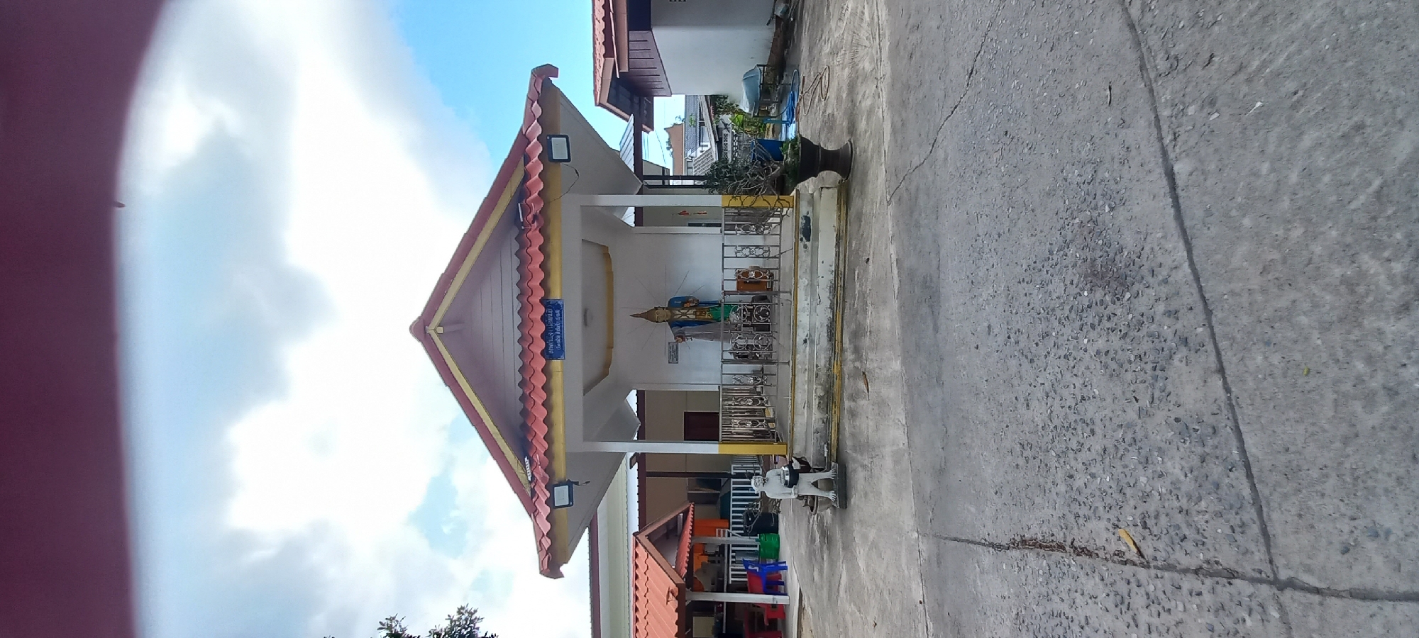 This media file captures a sunny outdoor scene in Baan Chaloklum, Thailand, featuring a traditional-style building next to a paved area. The main structure has a distinctive multi-tiered, gabled roof covered with red corrugated tiles and accented with yellow trim. White columns support the open-fronted porch area, under which two black floodlights are mounted.
Inside the porch, a prominent, colorful statue—primarily blue, gold, and green—stands on a raised platform, possibly a Buddhist deity or revered figure, positioned behind an ornate metal railing. Above the statue, a blue rectangular sign with unreadable white Thai script is affixed to the building's wall.
The foreground and right side of the image show a grey concrete or asphalt paved surface, marked with cracks and visible aggregate. On this paved area, near the building, there is a dark, decorative planter containing a small, gnarled plant. To the right of the planter, a stylized white statue of a standing figure is visible. In the background, other buildings with similar architectural elements can be glimpsed, along with some colorful plastic chairs. The sky is bright blue with scattered white clouds, indicating clear and sunny weather during daytime. No specific activity is taking place; the scene appears calm and static.