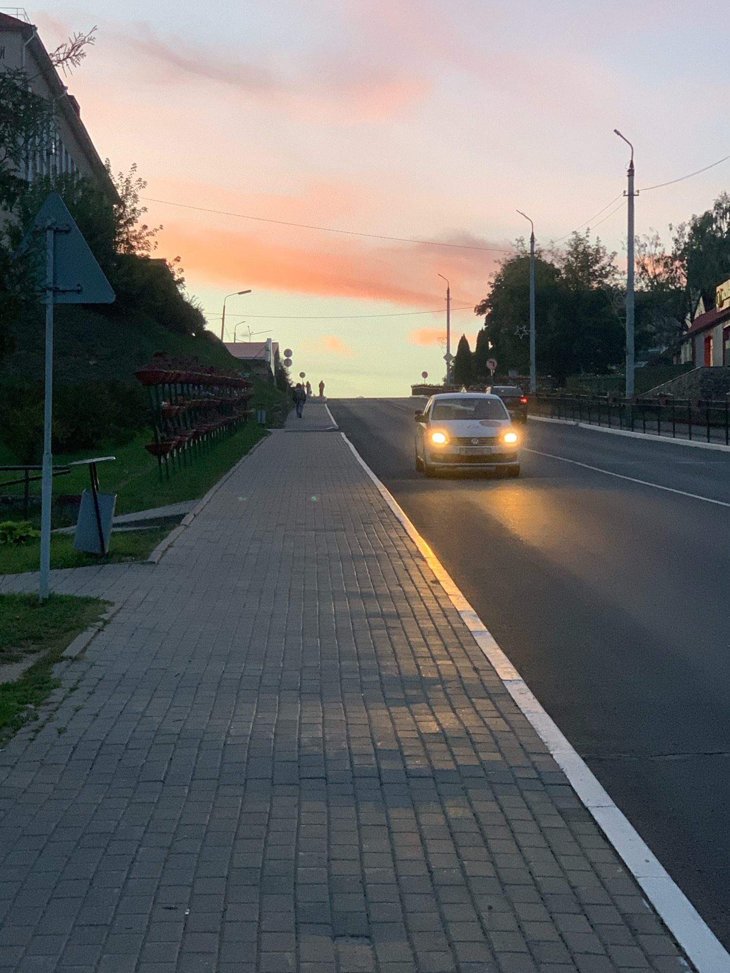The image captures a street scene in Kapyĺ, Belarus, during what appears to be twilight. A light-colored Volkswagen hatchback with its headlights on is driving uphill on an asphalt road on the right side of the frame. The road is bordered by a white line and a paved sidewalk made of grey rectangular blocks on the left. The sidewalk has a slight incline and is adjacent to a grassy embankment.
On the embankment, a series of dark planter boxes are arranged in rows. Above the embankment, a building with a slanted roof and trees are visible. A triangular road sign is positioned on a pole near the edge of the sidewalk. Further up the road, a few pedestrians are walking away from the viewer. The sky above is illuminated with soft pink and orange hues, indicating the presence of a sunset or sunrise. Several streetlights are visible along the roadside. The overall atmosphere suggests a peaceful, urban evening.