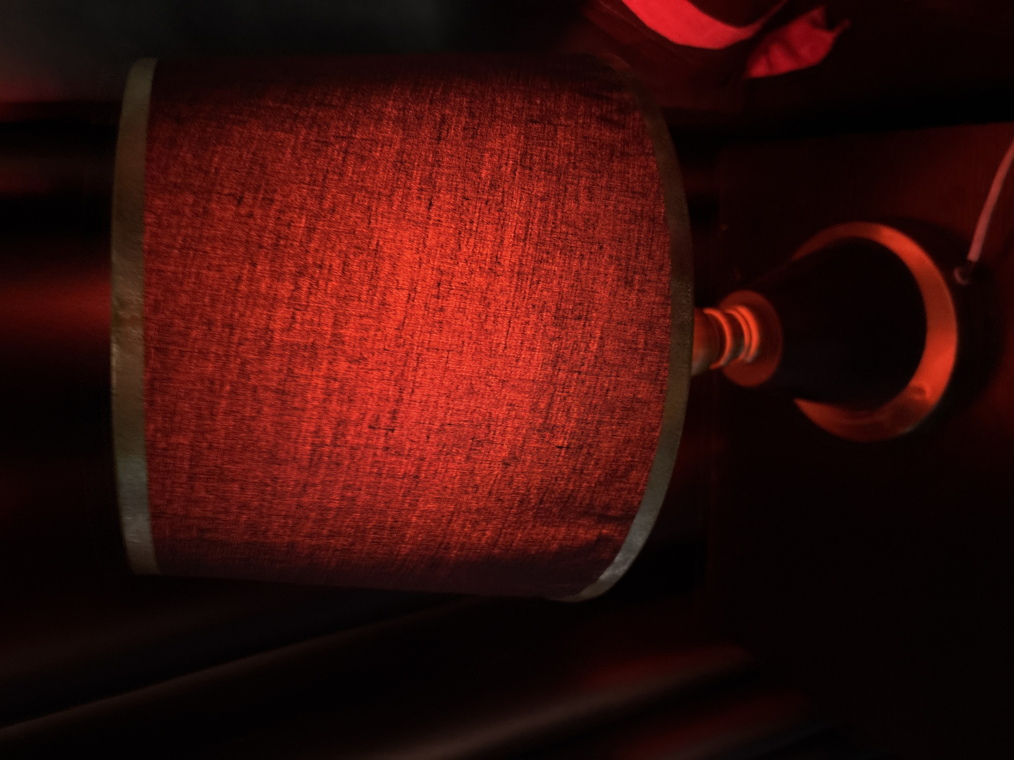 The image shows a close-up of a red lamp shade with a textured surface, lit from within, casting a warm red glow. The shade has a thin metallic trim along its top and bottom edges. Below the shade is the lamp base, partially visible, appearing to be made of dark material with metallic accents, one of which is illuminated by the red light. A white cable is plugged into a power source and extends from the base of the lamp. To the left of the lamp, dark, reddish-brown curtains are visible, suggesting a draped background. To the right of the lamp, another object with a red, curved surface is partially visible, possibly another piece of furniture or decor. The overall lighting is dim, with the red lamp being the primary light source, creating a moody atmosphere. This scene is located in the city of Obosi, Nigeria.