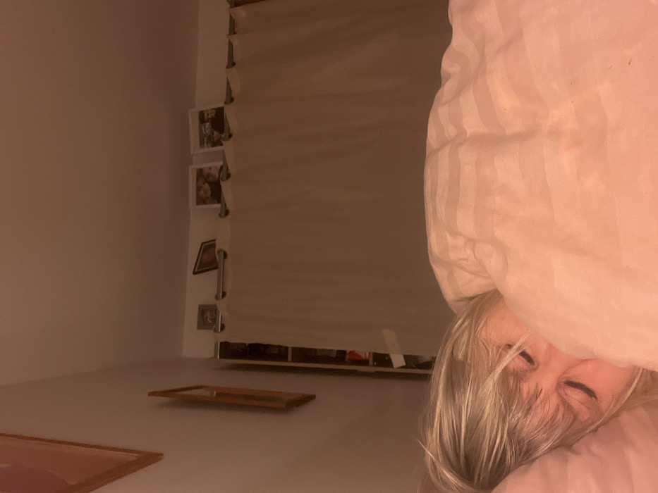 A person with blonde hair peeks out from under a pink striped blanket. Behind them, a window with drawn blinds is visible. Several framed photos are mounted on the wall beside the window. On the floor are two empty picture frames lying flat. The room is dimly lit, creating a soft, warm atmosphere.