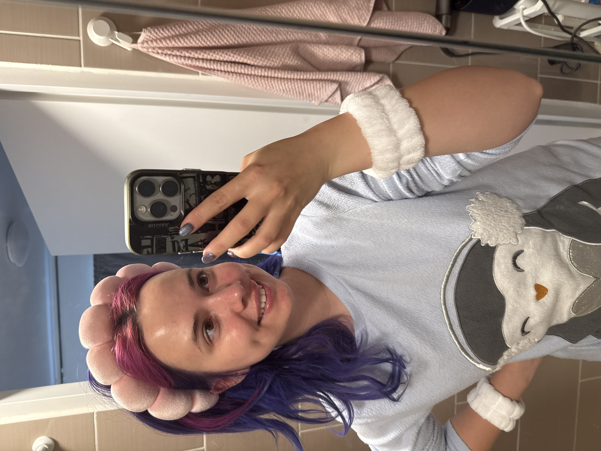 A young woman in Zagreb, Croatia, captures a mirror selfie, smiling brightly at the camera. She has striking two-toned hair, primarily purple with hints of magenta/pink. On her head, she wears a soft, light pink headband designed with rounded, petal-like elements, likely for skincare. She also has fluffy white wristbands on both arms, matching the soft texture of her light blue top, which features a large applique of a relaxed penguin-like character with closed eyes and an orange nose.
Her right hand holds a dark-cased smartphone, visible with its triple camera array and some obscured designs on the case. Her fingernails are painted with a dark, sparkly polish. The reflection of text "80 C00R" is faintly visible on the phone case.
The setting appears to be a bathroom, with light-colored (tan/beige) tiled walls and a white surface visible in the background. A light pink, waffle-weave towel hangs from a white suction cup hook on the tiled wall behind her. The overall impression is one of relaxation, possibly during a morning or evening routine, given her casual attire and the accessories.