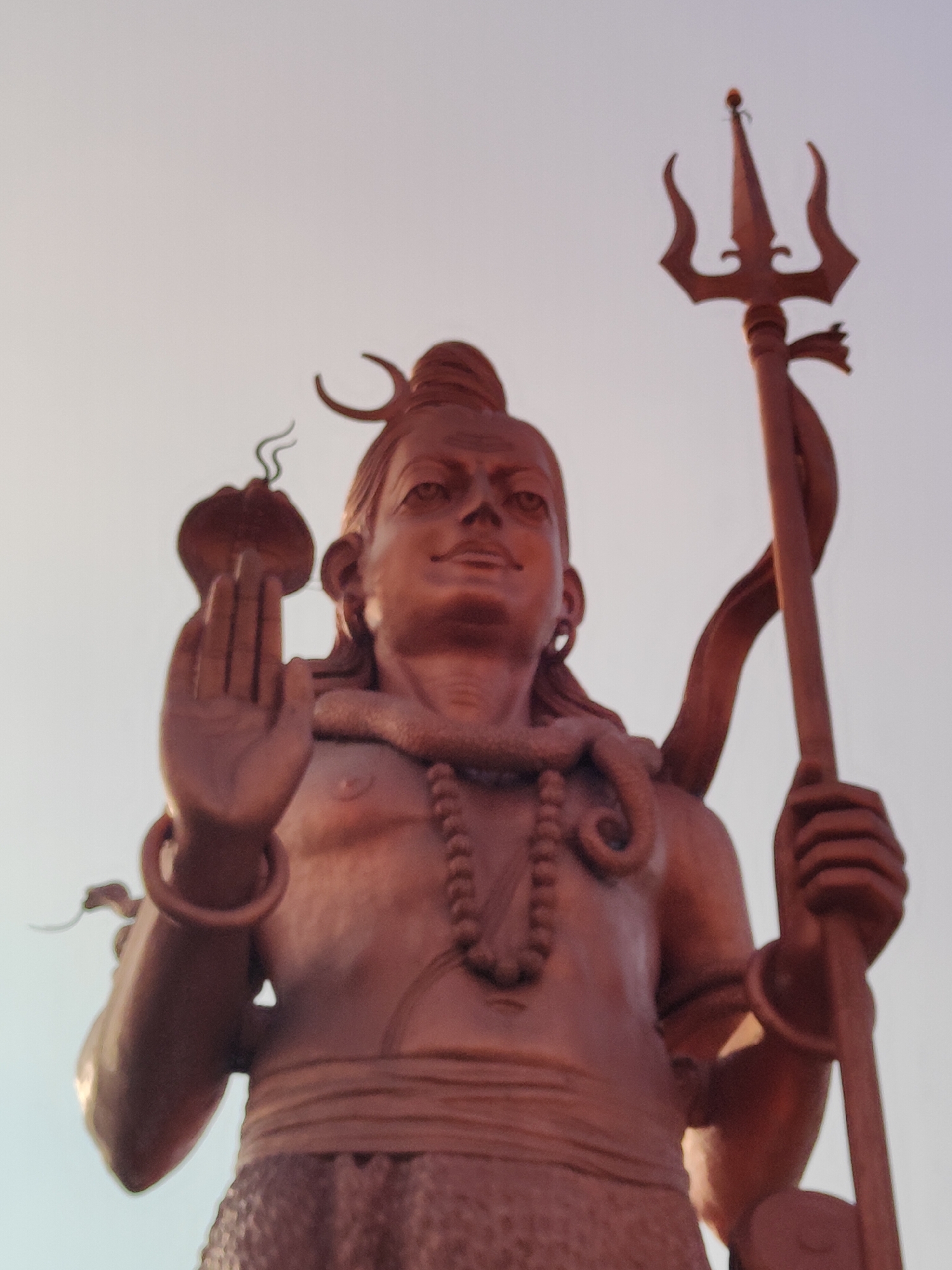 A low-angle, eye-level shot captures a bronze statue of Shiva, a Hindu deity, against a pale, overcast sky. The statue is predominantly reddish-brown, with a weathered patina. Shiva is depicted with his hair piled high and adorned with a serpent. He holds a trident, a symbolic weapon, in his right hand. His left hand is raised, palm outward, in a gesture of blessing or protection. A necklace of large beads adorns his chest, and another serpent is coiled around his neck. A sash or belt cinches his waist, above a patterned lower garment. The statue is located in the city of Gurgaon, India, specifically at the Sheetla Mata Temple, which is a prominent landmark. The lighting is soft and diffused, suggesting it is either early morning or late afternoon. The overall impression is one of grandeur and spiritual significance.