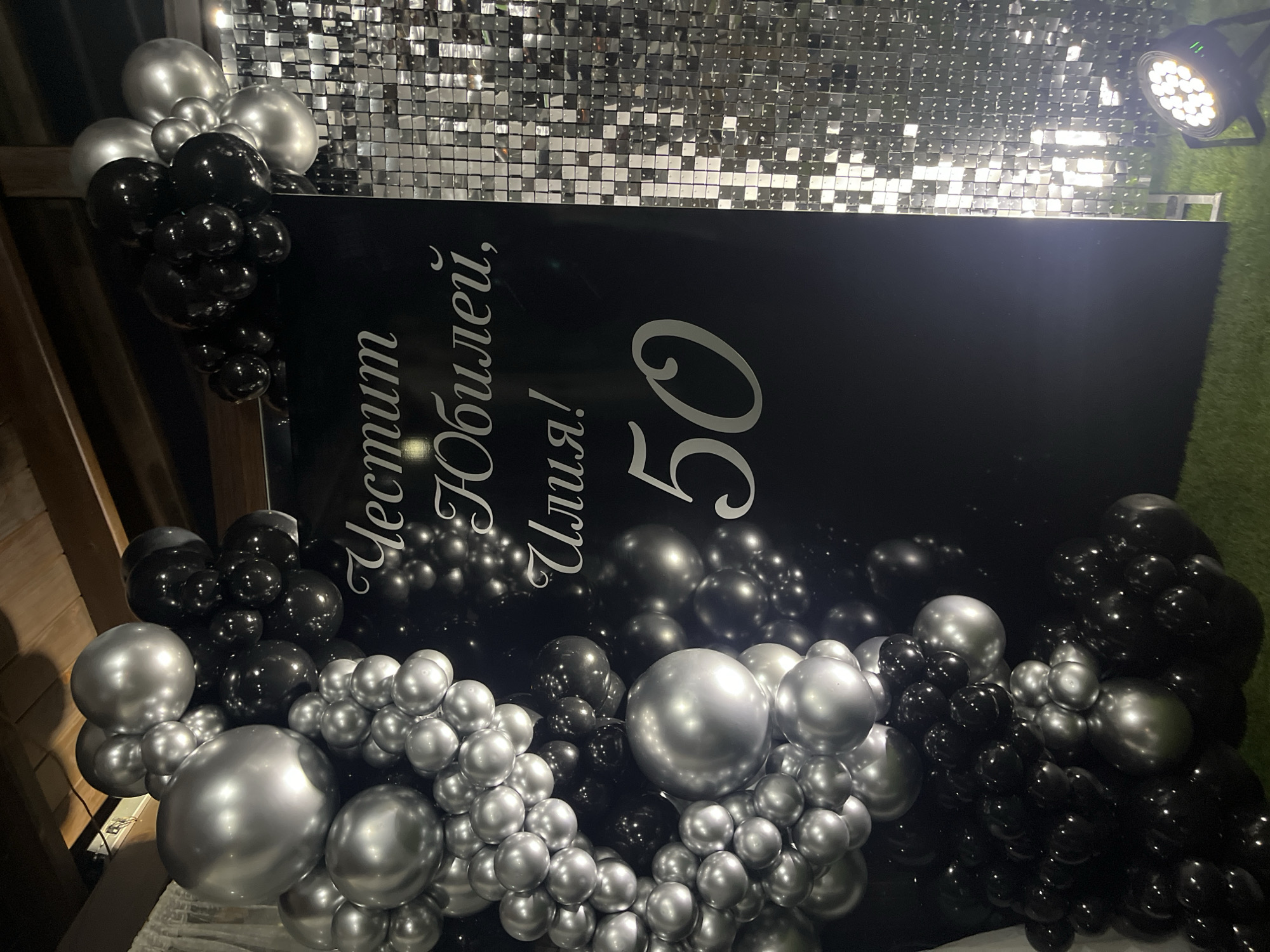 The image shows a black backdrop with silver text "Честит Юбилей, Илия! 50" (Happy Anniversary, Ilia! 50). The backdrop is decorated with an arrangement of black and silver balloons on the left and right sides. Behind the backdrop is a wall covered in small, square silver tiles that create a shimmery effect. In the upper right corner, there is a lighting fixture attached to what appears to be green artificial turf. The location is not discernible from the image. There is no information on whether this image was taken in Goce Delcev, Bulgaria.
