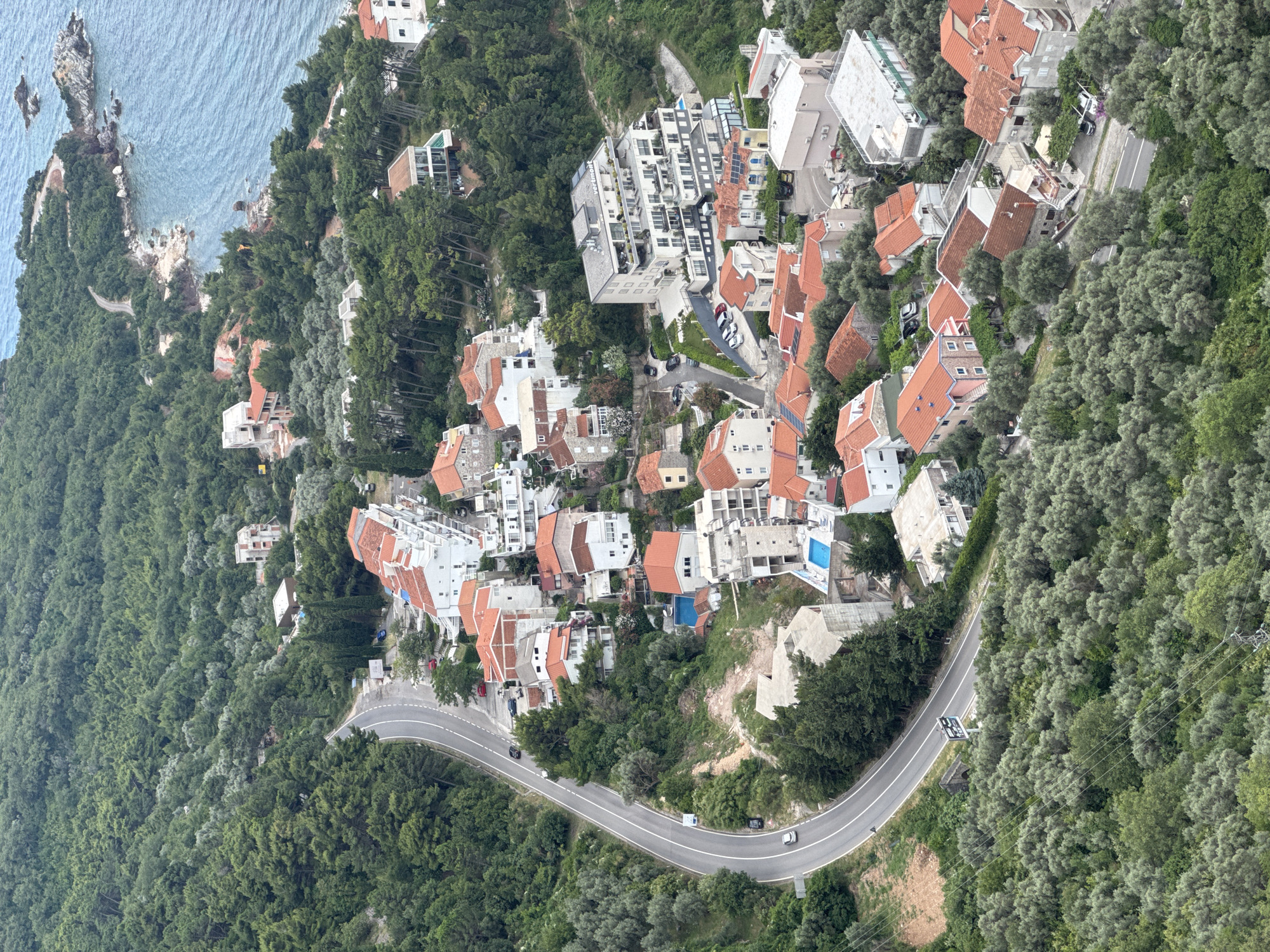 This is an aerial, landscape-oriented photograph capturing a coastal settlement nestled within a dense, green forested hillside. The scene is dominated by a winding road that curves through the trees, with a scattering of buildings visible on either side. The architecture primarily consists of multi-story structures with red-tiled roofs and white or light-colored facades, some featuring balconies and terraces. A few buildings appear more modern, with large glass windows.
The surrounding vegetation is lush and varied, with numerous trees creating a natural canopy over the landscape. The ocean is visible in the upper left portion of the image, its blue waters lapping against a rocky coastline with small islands. Several cars are present on the road, suggesting ongoing activity. Two swimming pools, one blue and one lighter, are visible on the rooftops of some buildings, indicating residential or hotel accommodations.
The time of day appears to be daytime, likely overcast or with diffused sunlight, as there are no strong shadows. The overall atmosphere is one of a peaceful, built-up area integrated into a natural, scenic environment. There is a road sign visible on the right side of the road with a speed limit displayed. The location is identified as Sveti Stefan, Montenegro.