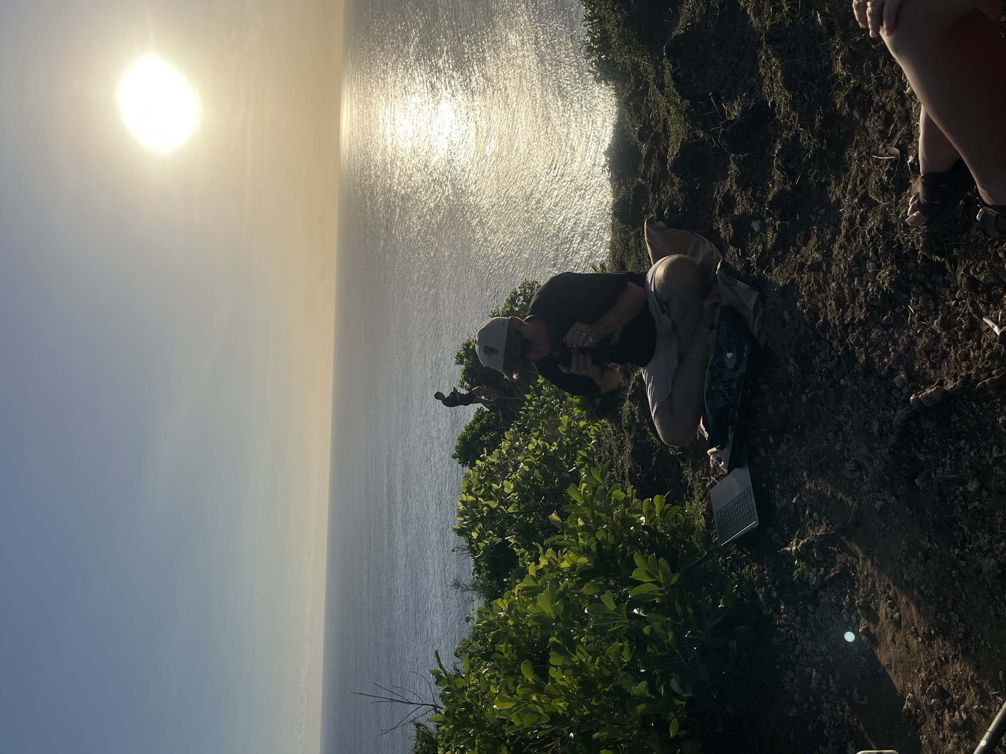 A high-angle, outdoor shot, rotated 90 degrees, shows two individuals on a cliff overlooking the ocean at Kuta Selatan, Indonesia. The sky is partly sunny with the sun positioned in the upper left corner. The ocean, which occupies a significant portion of the background, reflects sunlight. One individual, dressed in a dark shirt and light-colored shorts and wearing a baseball cap, is kneeling on the edge of the cliff, seemingly looking at or interacting with something in front of them. Beside them is a laptop on the ground. Another person is cropped on the upper right of the image. The edge of the cliff is covered with green vegetation and patches of bare earth. In the mid-ground, a single person can be seen standing at the edge of the cliff facing the ocean.
