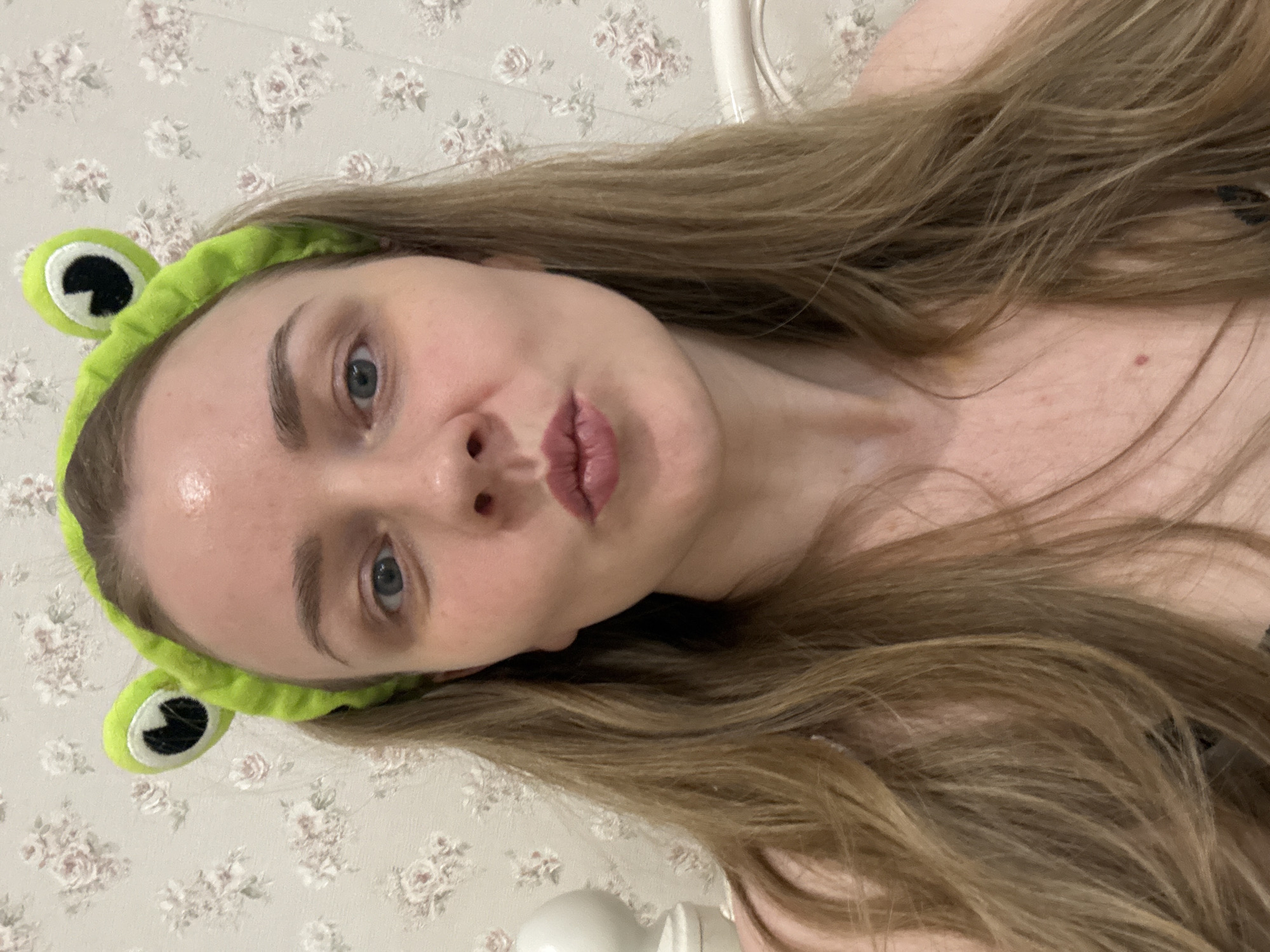 This close-up portrait features a young woman, likely in her 20s or 30s, situated indoors in Chaikovsky, Russia. Her face is directed towards the camera, with light blue or grey eyes looking straight ahead and her lips slightly pursed in a pouting expression, conveying a playful or contemplative mood. She has fair skin, which shows a subtle, healthy sheen on her forehead. Her long, wavy hair, a shade of light brown or dirty blonde, flows down around her shoulders. Adorning her head is a distinctive bright green headband designed with protruding, cartoonish frog eyes, positioned near her temples. The background is a light-colored wallpaper patterned with delicate, muted floral designs, primarily depicting roses in shades of pink and green. A portion of a white, curved object, possibly a bed frame or furniture detail, is visible in the upper right corner, suggesting a bedroom setting. No visible text is present in the image. The composition is a tilted, intimate shot, likely a selfie.