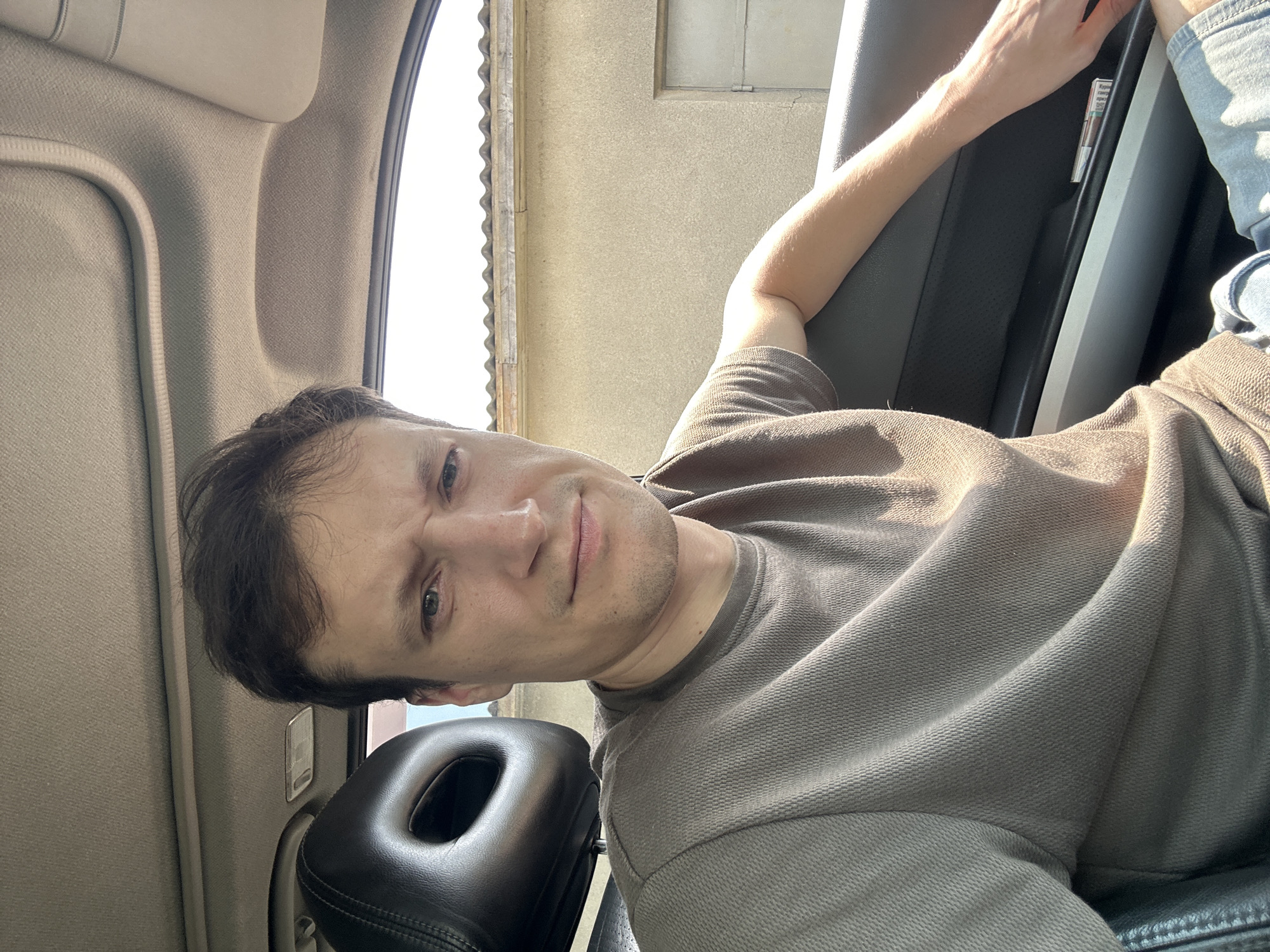 A man with short brown hair and a scowl sits in the driver's seat of a car. He is wearing a brown t-shirt. The car interior, including a black leather headrest and door panel, is visible. Outside the car, a tan building with a corrugated metal roof can be seen. The lighting suggests it is daytime. The exact location in Shatava, Ukraine cannot be determined from the visual information provided.