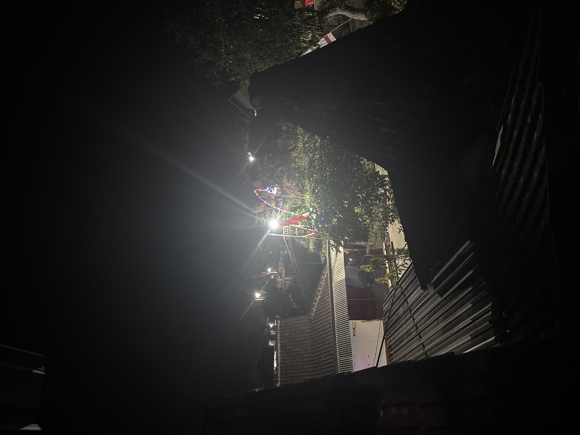 The image is a nighttime exterior shot taken from an elevated perspective, looking over rooftops in the village of Tirtomoyo, Indonesia. The foreground is dominated by the dark, silhouetted shapes of two adjacent rooftops. The rooftop on the left is a dark, corrugated metal structure, angled towards the viewer. To the right, a more prominent, dark, pointed structure, possibly the peak of a roof or an attic space, also obscures the view.
Beyond the immediate rooftops, the scene opens up to a dimly lit village landscape. Several houses with tiled roofs are visible, with one building featuring a white exterior and red accents. The area is illuminated by streetlights that cast long, bright beams into the night sky, creating lens flares. In the mid-ground, a cluster of trees and foliage are illuminated, some of which appear to have festive lights strung through them. Several red and white flags, consistent with the Indonesian flag, are prominently displayed on poles, suggesting a celebration or national holiday. The background shows more distant structures and trees, fading into the darkness of the night. No people are discernible in the image.