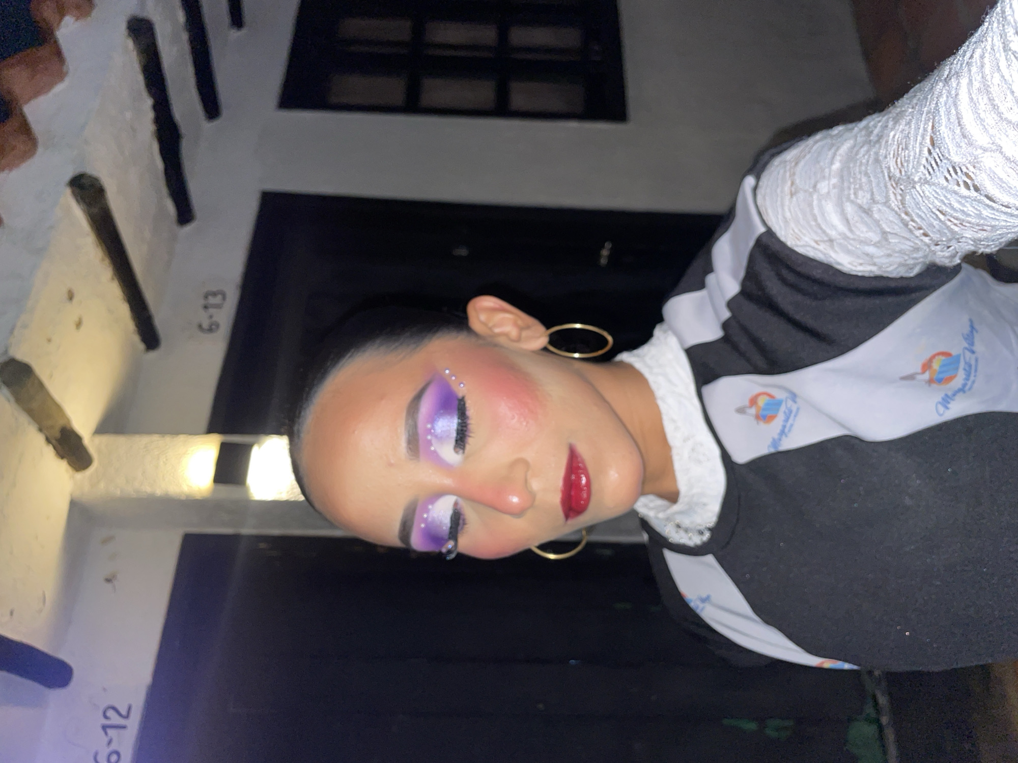 A young woman with dramatic purple eyeshadow and glittery accents is pictured outdoors, her eyes closed. She wears a black and white top with a white lace collar and sleeves. The background shows a building with numbered doors or units visible (6-12 and 6-13). The lighting suggests it is evening. The overall impression is of a staged portrait, possibly before or after a performance or event, given the elaborate makeup and costume details. The presence of the building numbers suggests a residential complex or a series of small businesses. The vegetation visible on the left side indicates an outdoor setting.