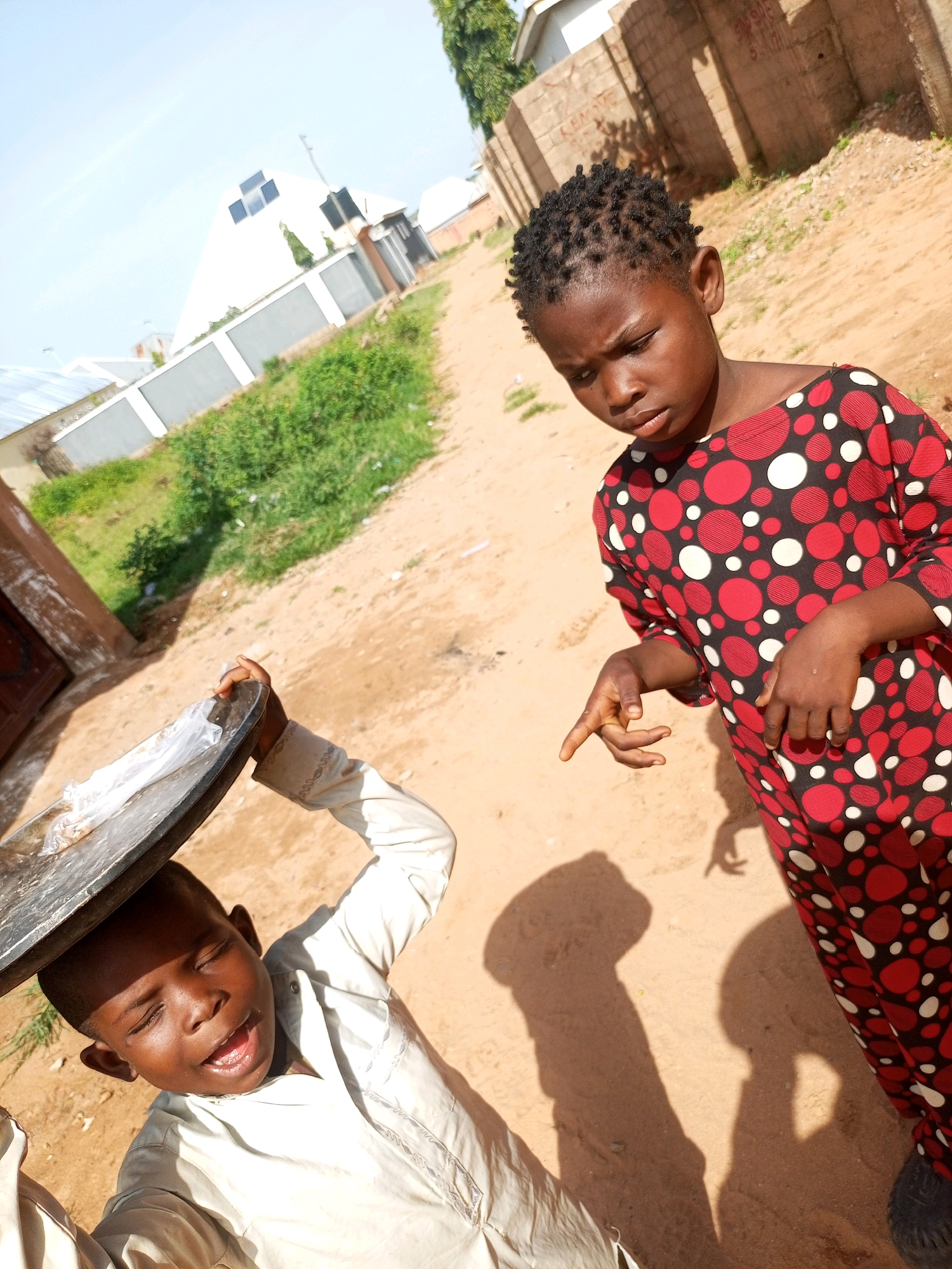 In Tempure, Nigeria, two children are standing on a dirt path. The child on the left, wearing a light-colored tunic, is holding a large metal tray above their head. Their eyes are closed, and their mouth is open, as if they are exerting effort or singing. The child on the right, wearing a red and black polka-dot dress, is looking down and extending their right hand with two fingers pointed. In the background, there is a white building with a triangular roof and solar panels. A brick wall and some trees are also visible. The ground is sandy and uneven, with some patches of dry grass. The overall lighting suggests it is a sunny day.