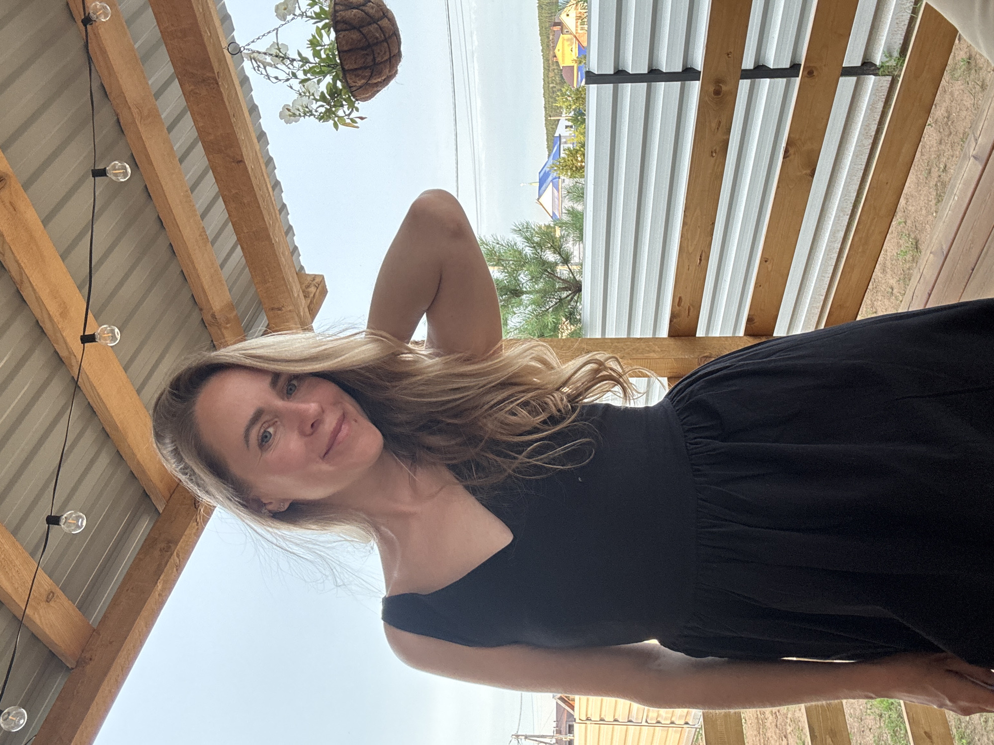 A woman with blonde hair and a black dress stands on a wooden porch. She is looking at the camera and smiling. Behind her, there is a fence and some trees. The sky is cloudy. This image was taken in Chaikovsky, Russia.