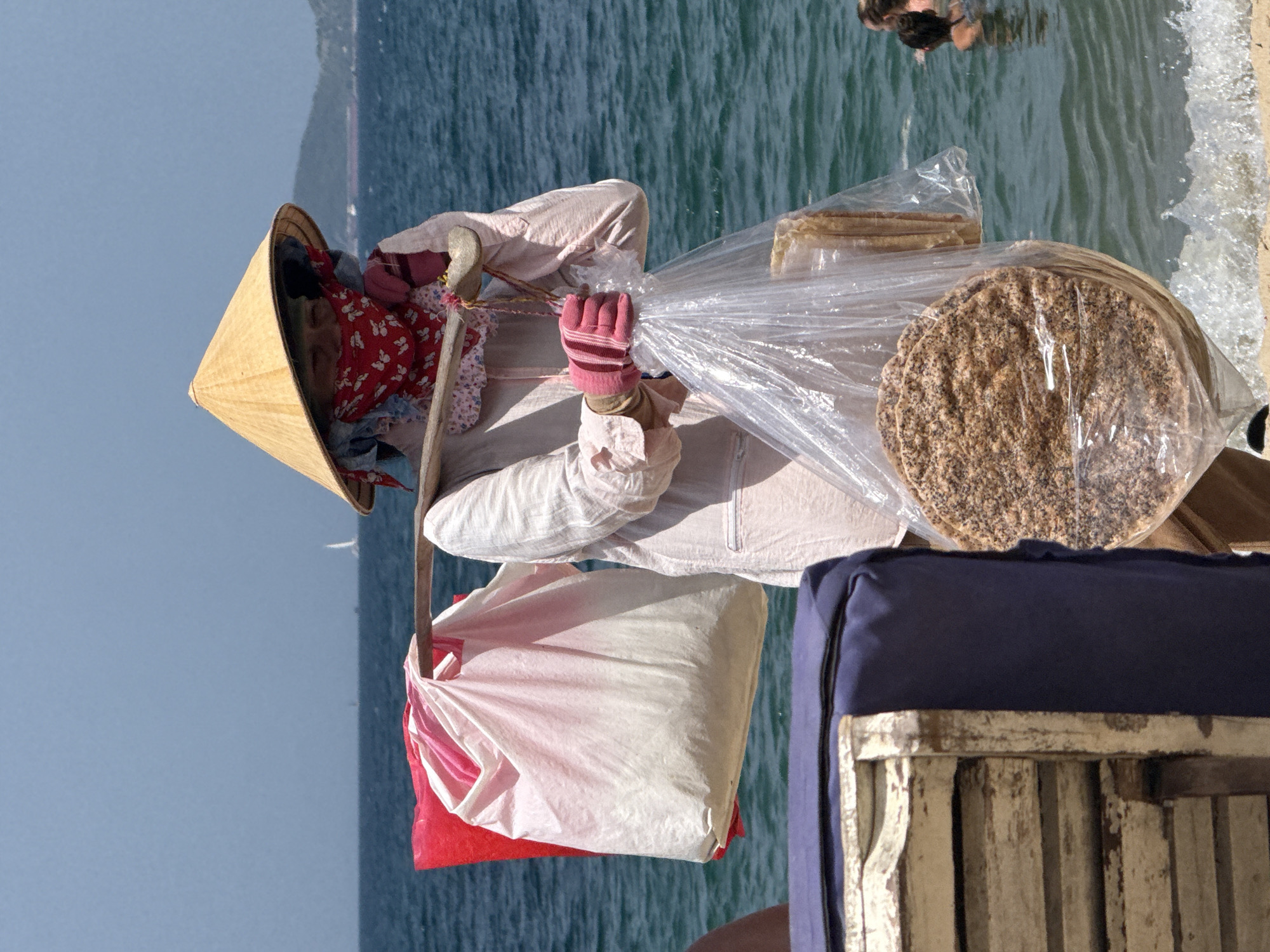 A person wearing a conical hat and a red bandana over their face carries goods suspended from a shoulder yoke in Nha Trang, Vietnam. The person is dressed in a light-colored, loose-fitting garment and pink gloves. They carry two large bags from the yoke, one red and white, the other a clear plastic bag containing round, flat food items. In the background, the turquoise ocean stretches to the horizon, with a faint sailboat visible. To the right, people swim in the water. In the foreground, a portion of a beach lounge chair with a dark blue cushion is visible. The scene captures a vendor on a beach, likely selling snacks to beachgoers.
