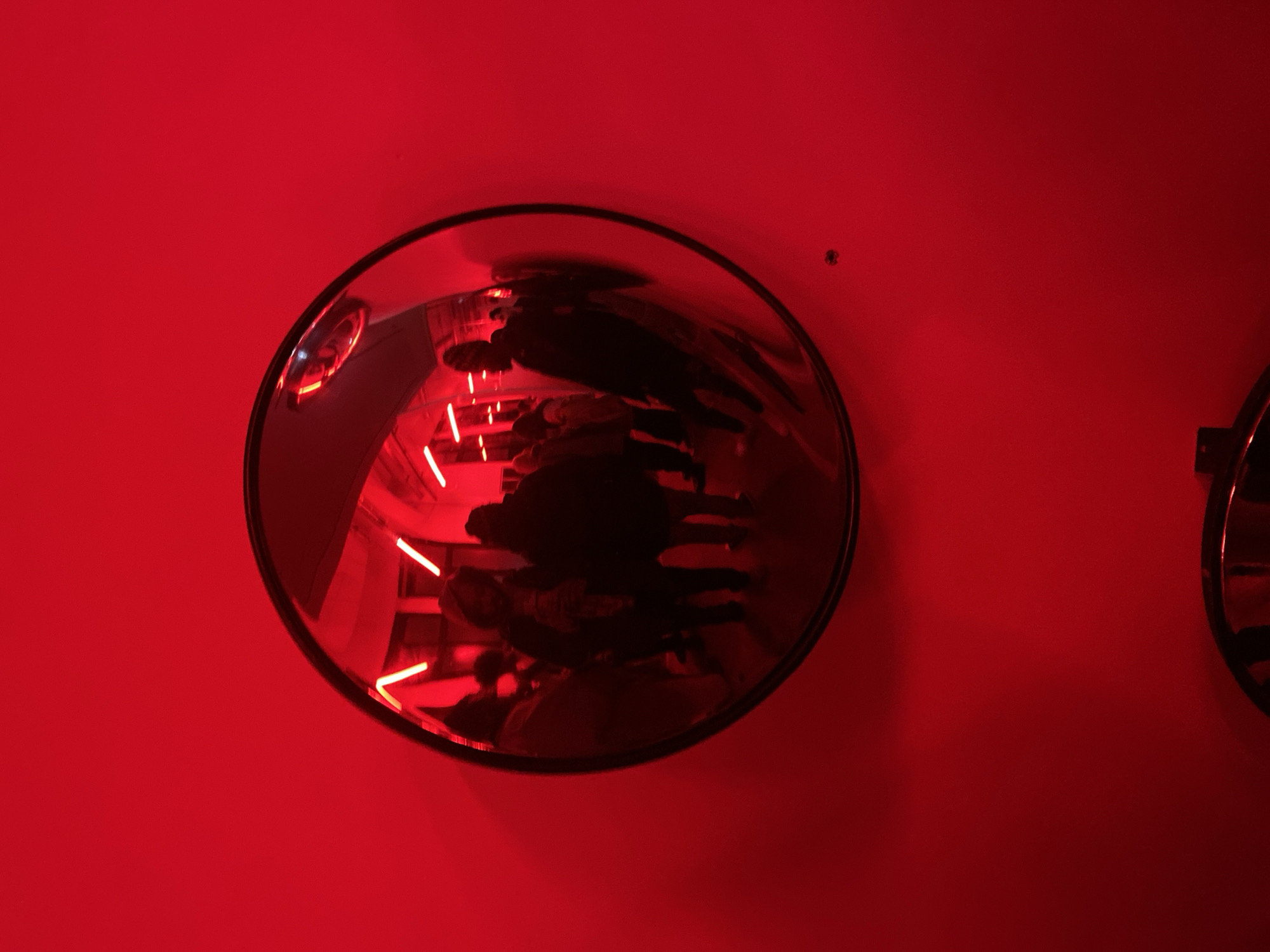 A surveillance mirror is mounted on a red wall in an interior space. The mirror reflects a red-lit room with people standing in a line. A second surveillance mirror is partially visible to the right.