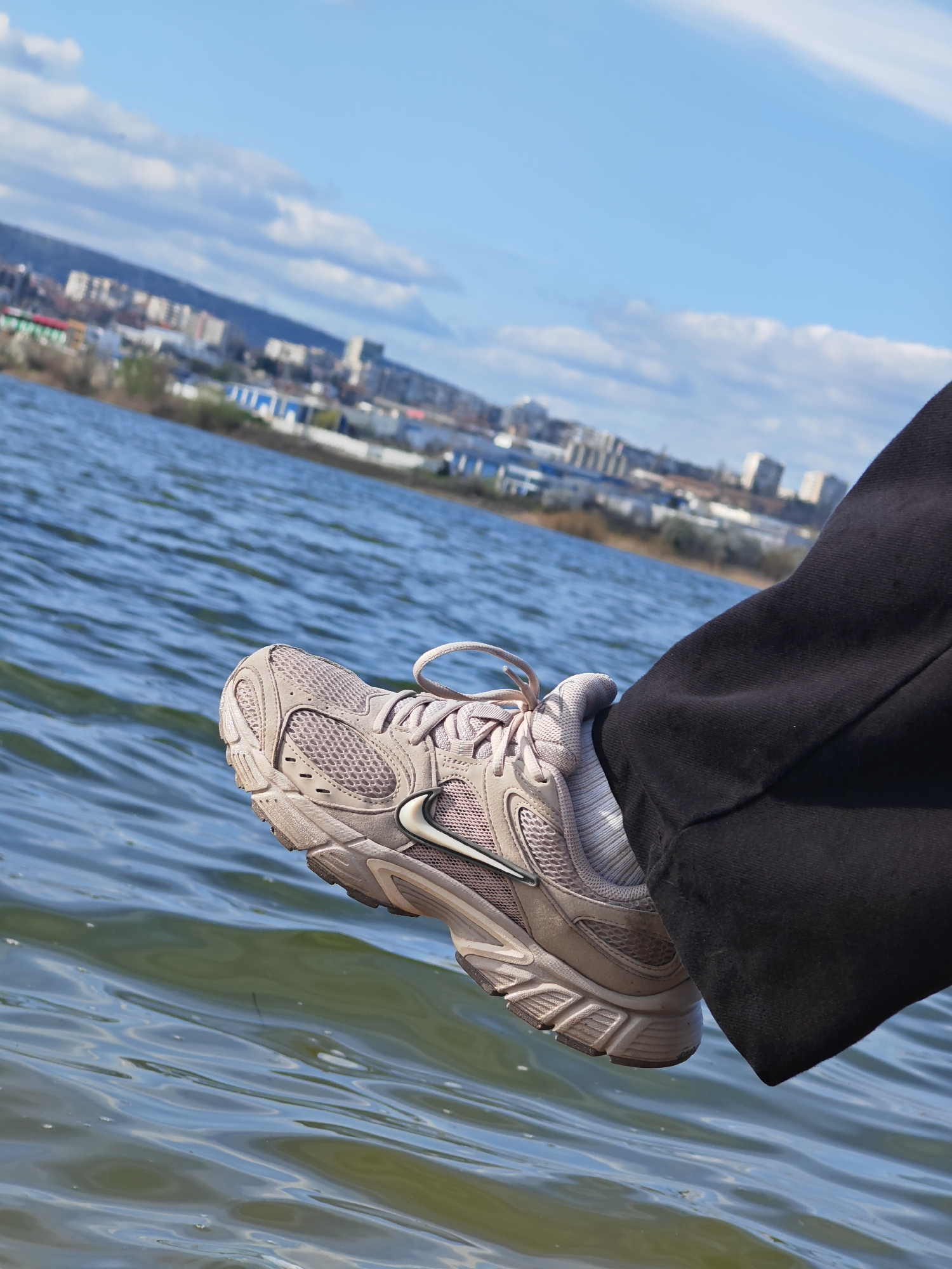The image shows a right leg wearing a dark-colored sweatpant and a light-colored Nike running shoe dangling over water. The water appears to be a body of water, possibly a lake or river, with ripples on its surface. Behind the water, there is a cityscape under a blue sky with scattered clouds. The buildings are a mix of residential and industrial structures. Based on the cityscape and confirmed by online search, the location is in the city of Shumen, Bulgaria, likely near the artificial lake of Kyoshkovets. The person is not visible, only the leg is within the shot. The action is dangling a foot over the water.